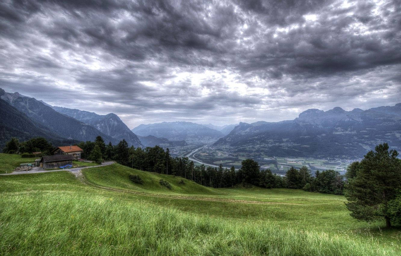 Liechtenstein 桌布 壁紙 — 一個戲劇性的山谷，有茂密的綠色草地，在厚重的陰天下，遠處有山峰和一間小屋。