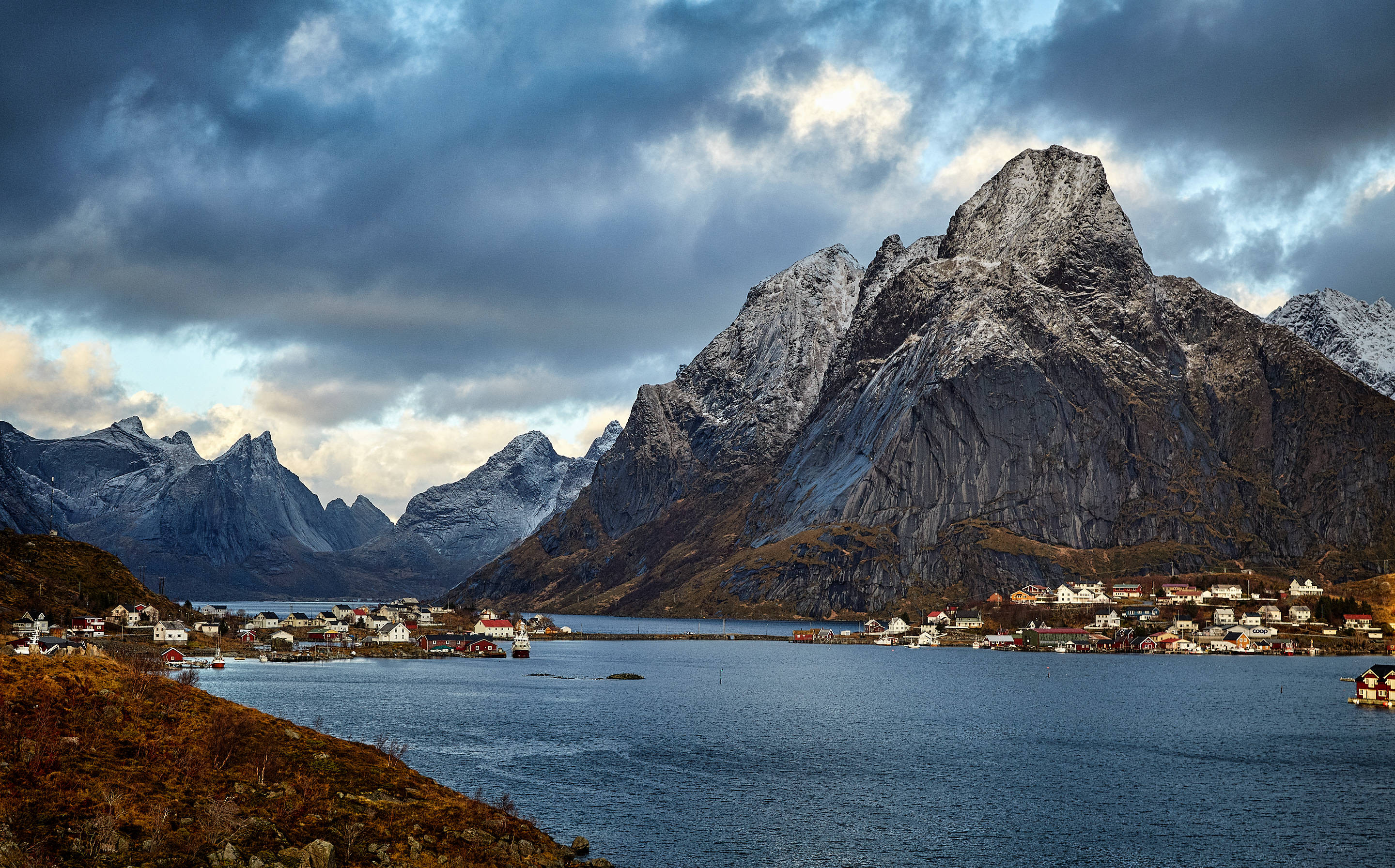 Norway 桌布 壁紙 — 一個戲劇性的峽灣村莊，坐落在白雪覆蓋的山脈之間，天空陰沉多雲。