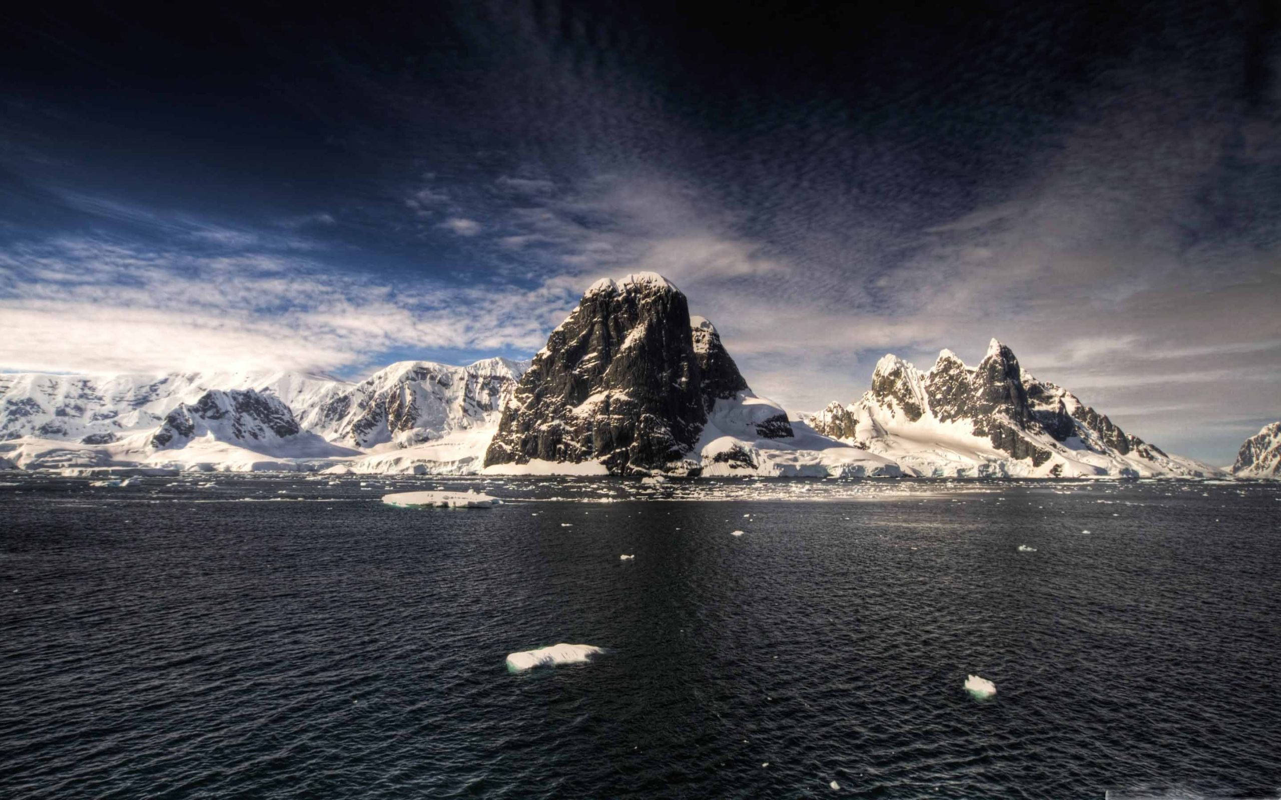Antarctica 桌布 壁紙 — 一個戲劇性的極地景觀，白雪覆蓋的鋸齒狀山脈從點綴著浮冰的黑暗水域中升起，天空陰沉。