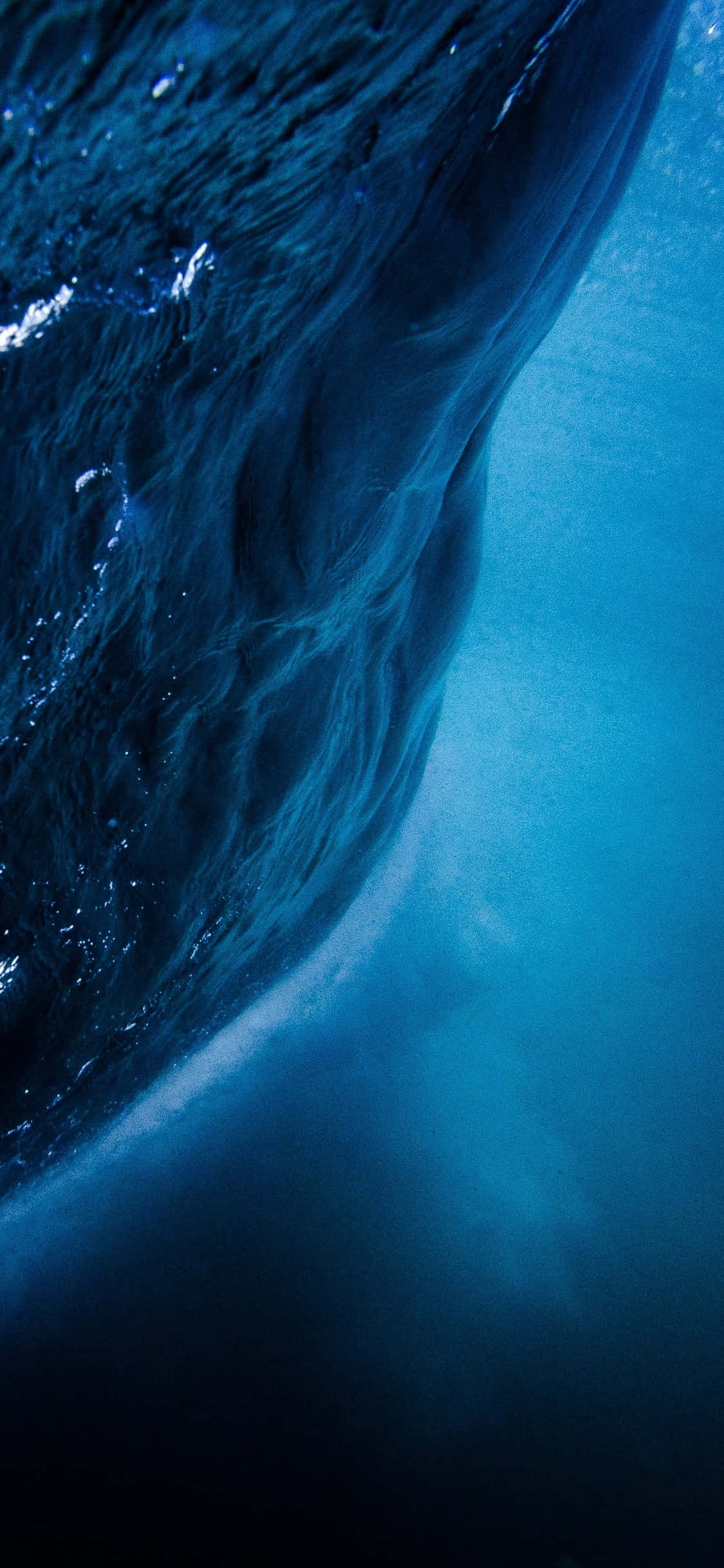 Underwater iPhone 桌布 壁紙 — 一個戲劇性的水下視角，巨大的海浪在頭頂捲曲，帶有深藍色紋理和光線穿過水體過濾。