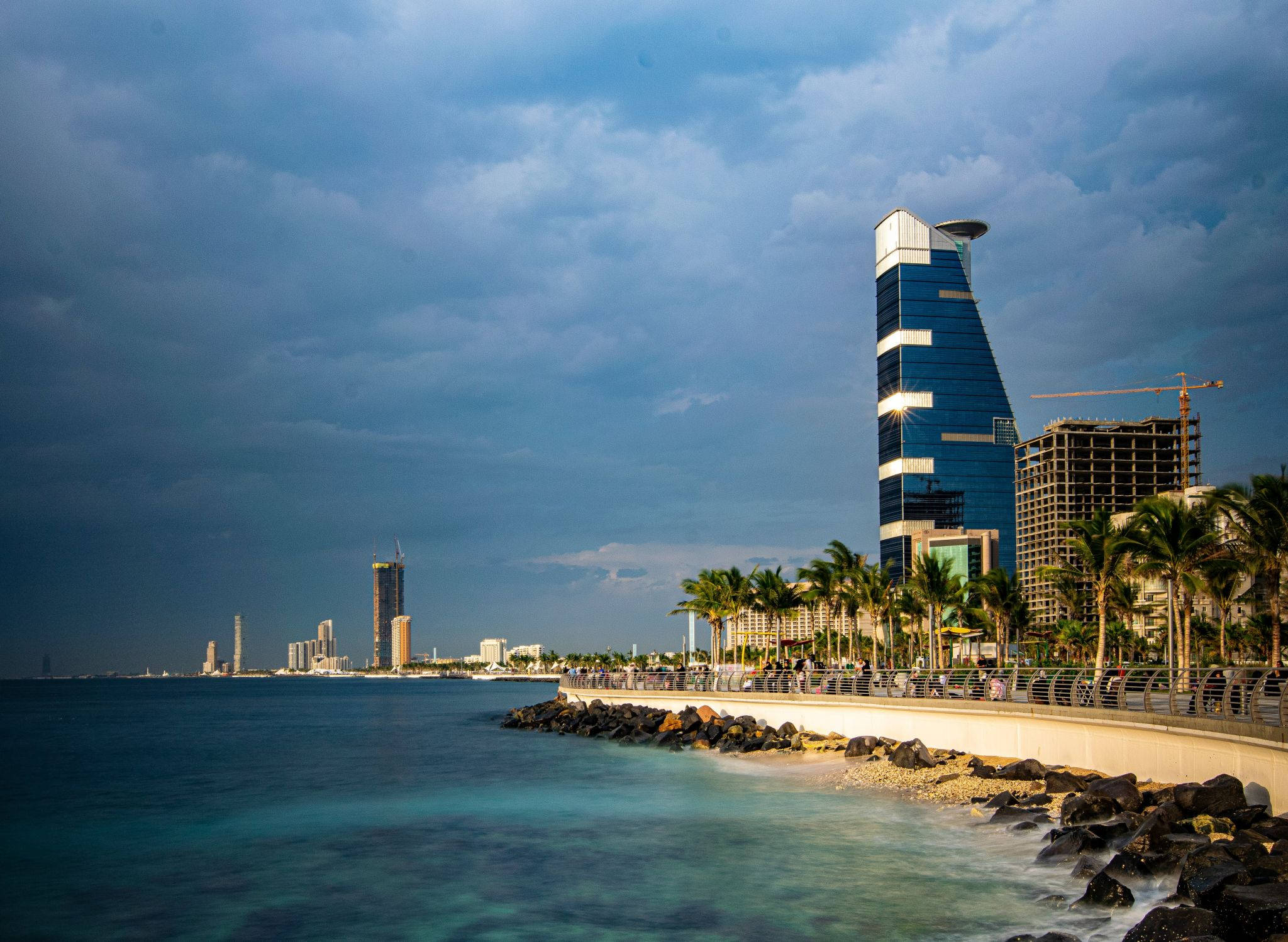 Saudi 桌布 壁紙 — 一個戲劇性的沿海城市景觀，在暴風雨的天空下有一座引人注目的藍色玻璃摩天大樓。