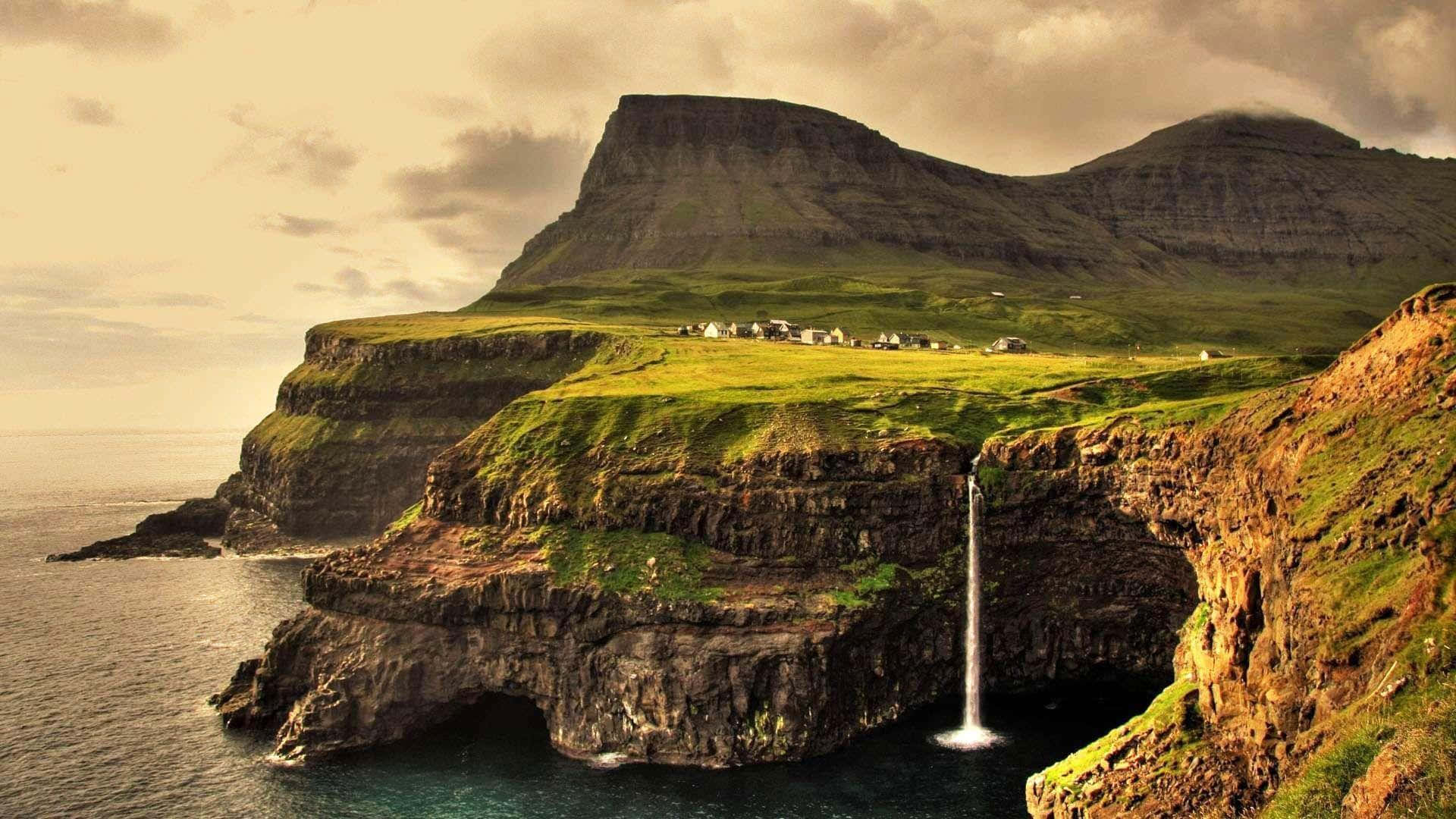 Aesthetic Nature 桌布 壁紙 — 一個戲劇性的海岸懸崖，瀑布從一個長滿草的高原傾瀉入海，高原上散落著小房子，天空多雲。