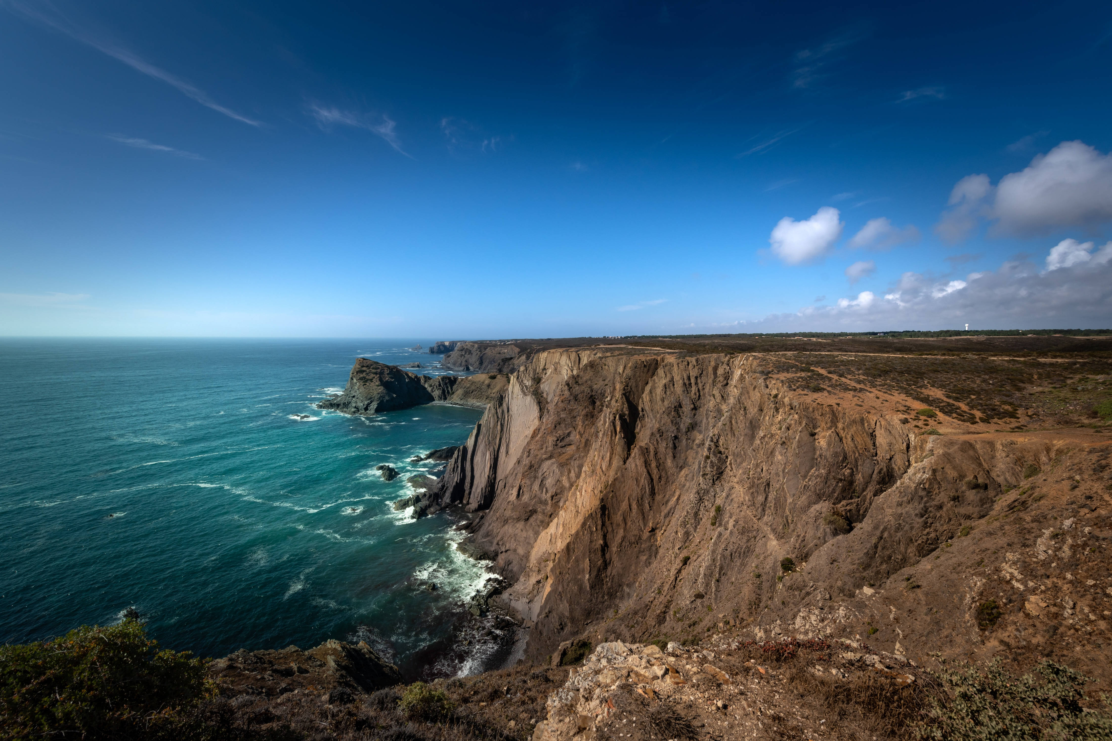 Portugal 桌布 壁紙 — 一個戲劇性的海岸懸崖墜入碧綠色的海浪中，在廣闊的藍天下有散落的雲朵。