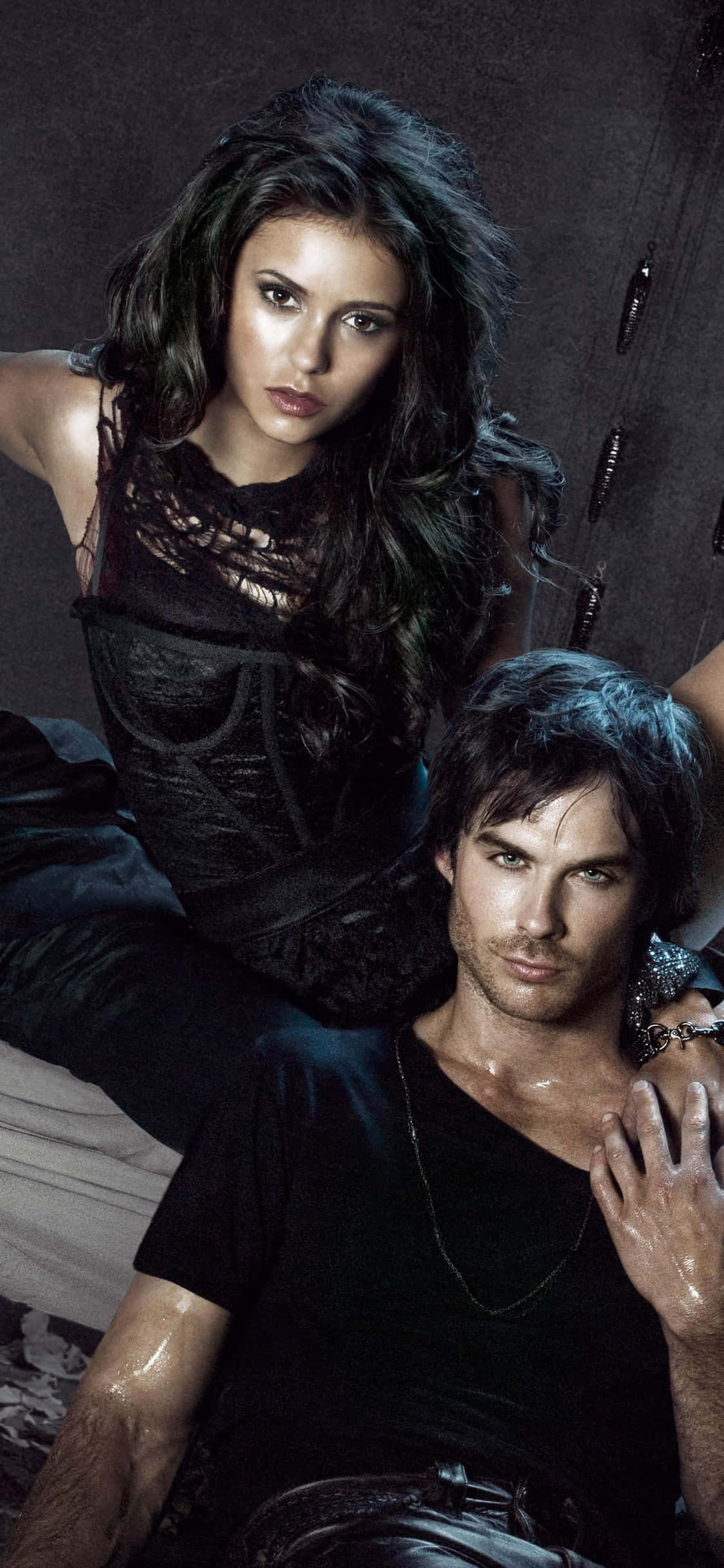 The Vampire Diaries iPhone 桌布 壁紙 — 一個戲劇性的肖像，一男一女穿著深色濕衣服，在陰影紋理牆前強烈地擺姿勢。