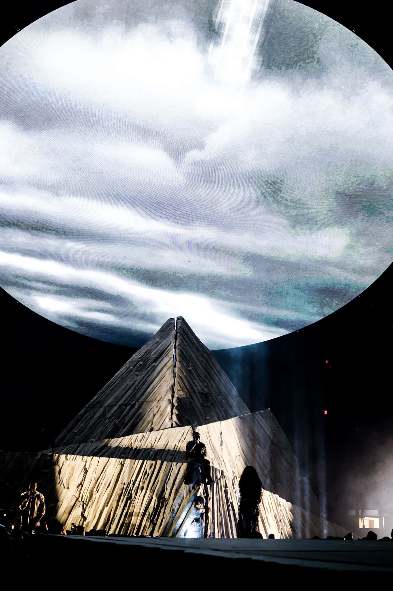 Kanye West Yeezus 桌布 壁紙 — 一個戲劇性的舞台，在巨大的圓形螢幕下有一個木製金字塔結構，顯示旋轉的雲和光束。