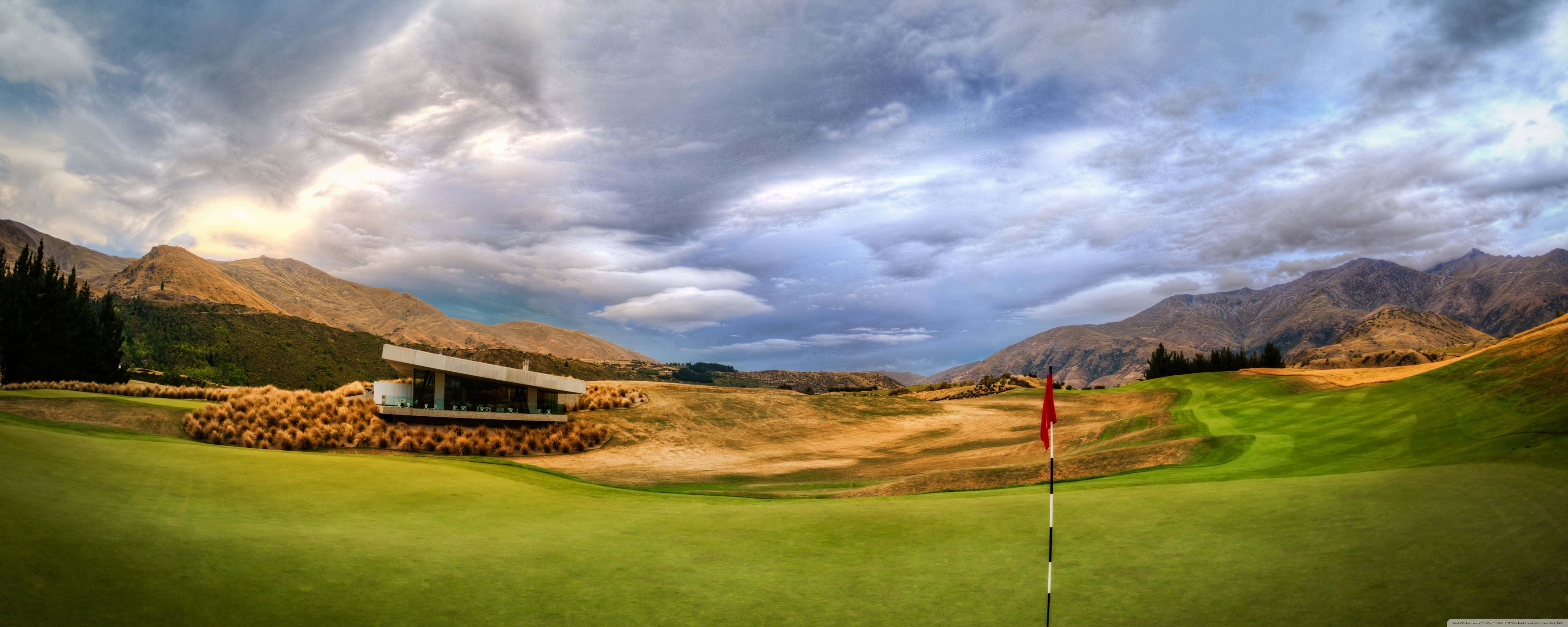 Golf 3840x1080 桌布 壁紙 — 一個戲劇性的高爾夫球場，果嶺上插著紅旗，周圍環繞著起伏的丘陵和多雲的天空。