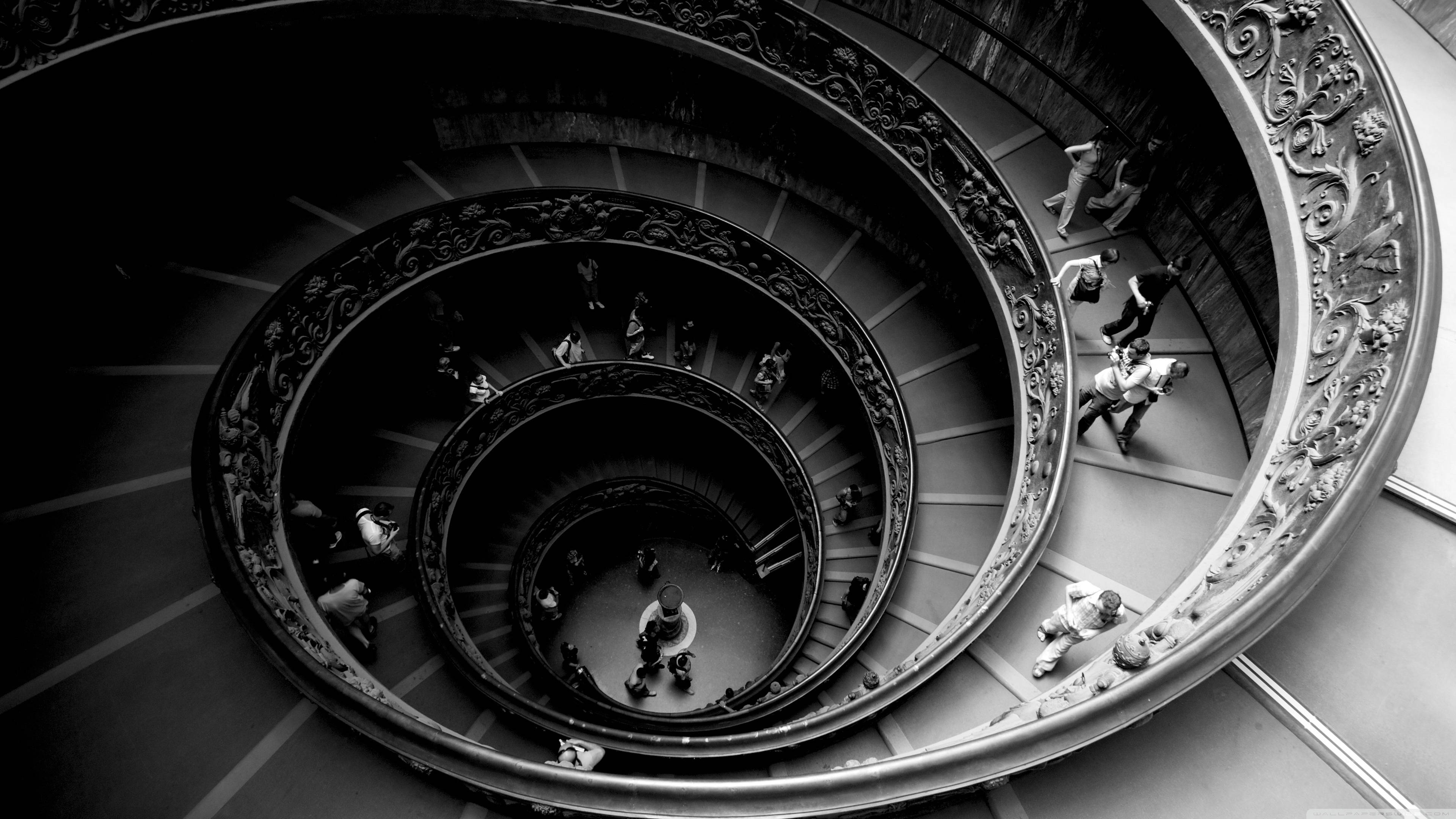 Spiral 桌布 壁紙 — 一個戲劇性的黑白俯瞰圖，展示了一個帶有華麗欄杆的宏偉螺旋樓梯，人們正在上下走動。