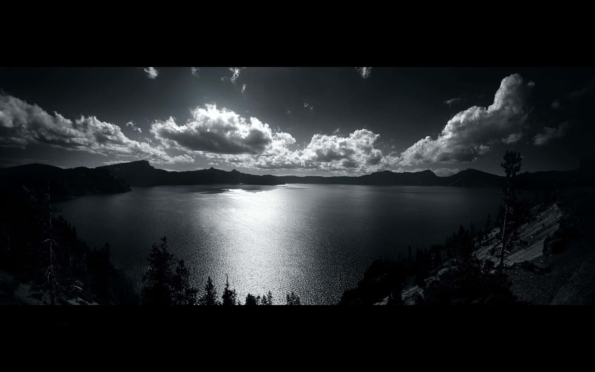 Dark Landscape 桌布 壁紙 — 一個戲劇性的黑白全景，展現廣闊湖泊透過暴風雲反射陽光，襯托出樹木剪影和遠山。