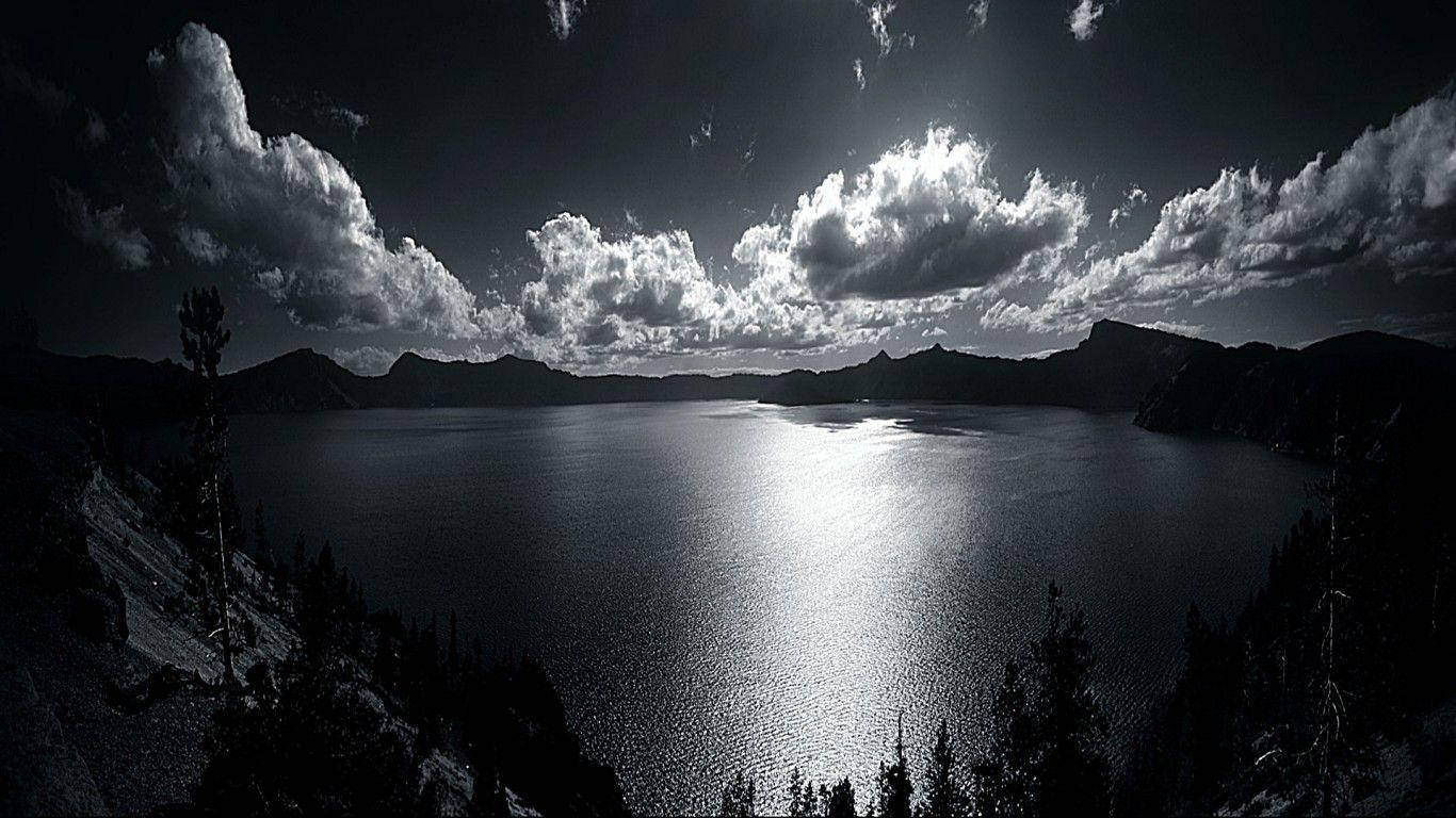 Lake 桌布 壁紙 — 一個戲劇性的黑白景觀，廣闊的火山口湖反射陽光穿過高聳的雲層，剪影樹木框住場景。
