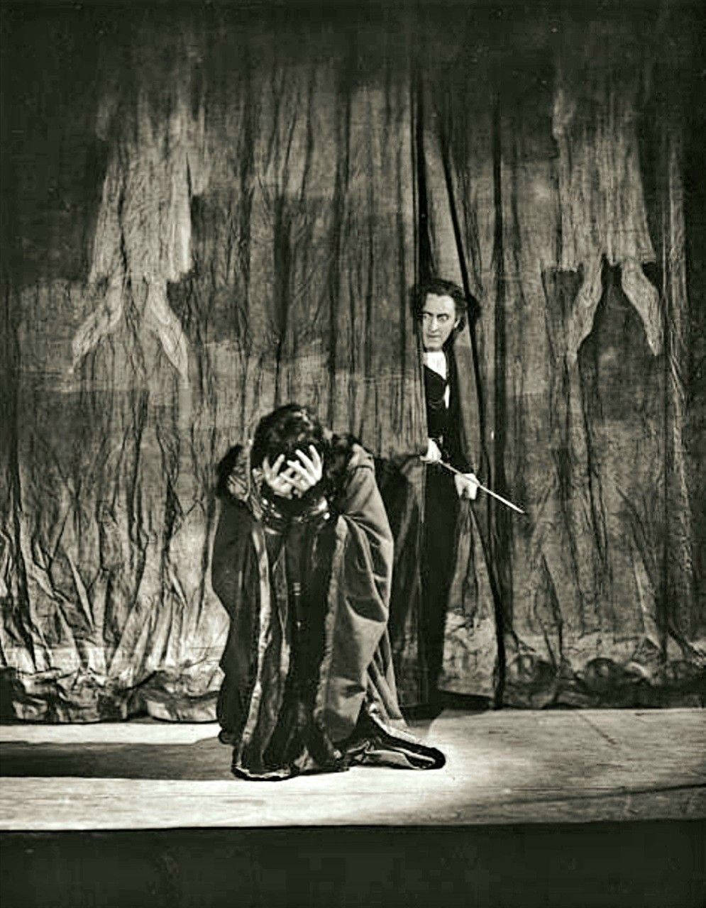 Hamlet 桌布 壁紙 — 一個戲劇性的黑白舞台場景，一名演員痛苦地跪著，而另一名演員站在厚重的窗簾後面，帶有威脅性。