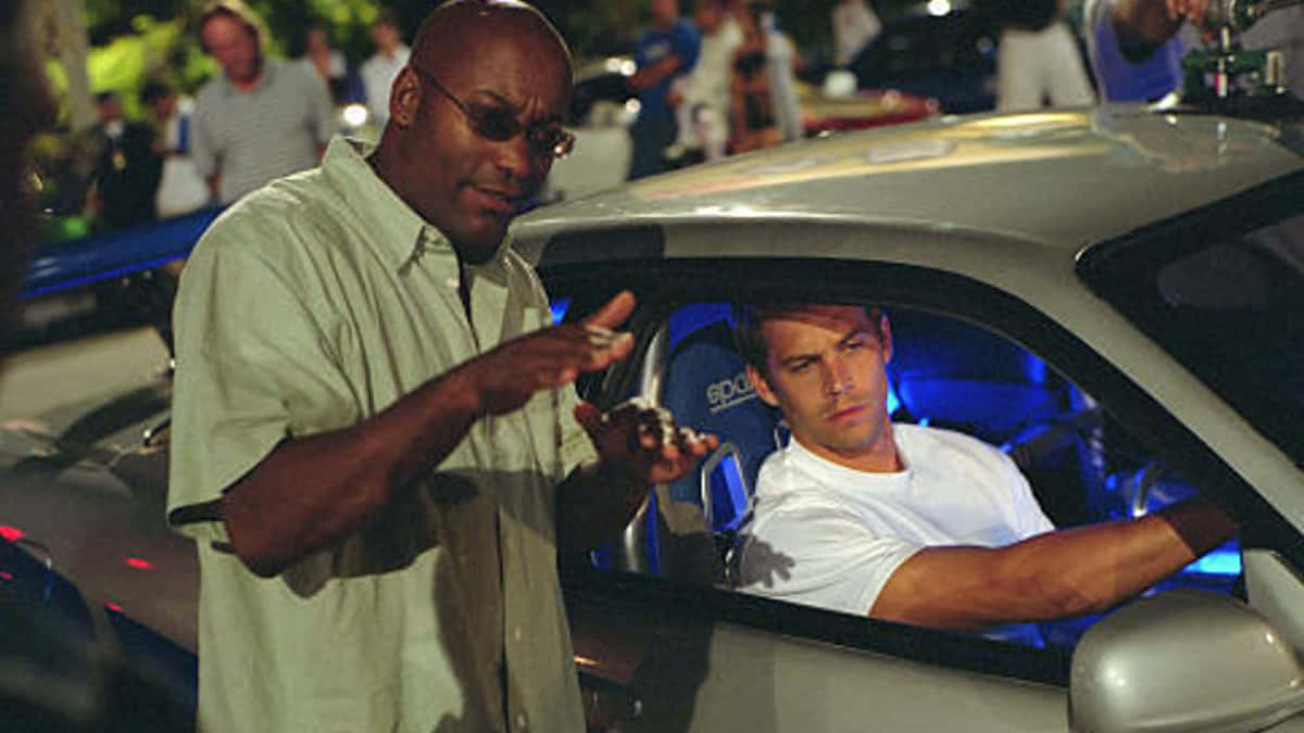 Paul Walker Skyline 桌布 壁紙 — 一個戴太陽眼鏡的男人急切地向另一個穿著白色T恤、坐在銀色跑車內的男人做手勢，背景是夜晚。