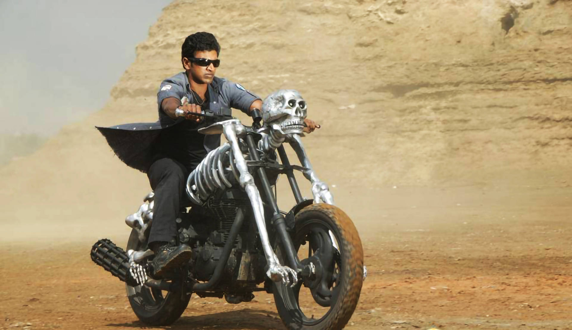Puneeth Rajkumar 桌布 壁紙 — 一個戴太陽眼鏡的男人騎著骷髏主題機車穿過塵土飛揚的沙漠景觀。