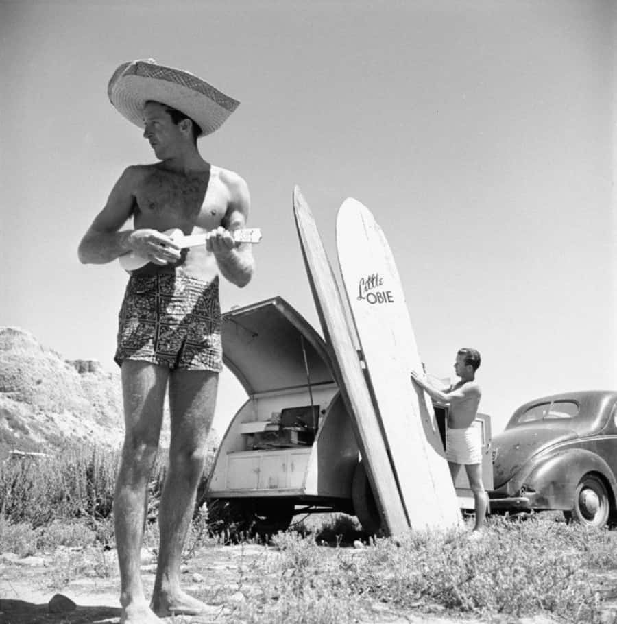 surfing 桌布 壁紙 — 一個戴寬邊帽的男人在復古露營車和衝浪板旁邊彈奏烏克麗麗，而另一個男人在陽光明媚的沙漠景觀中靠在一輛經典汽車上。
