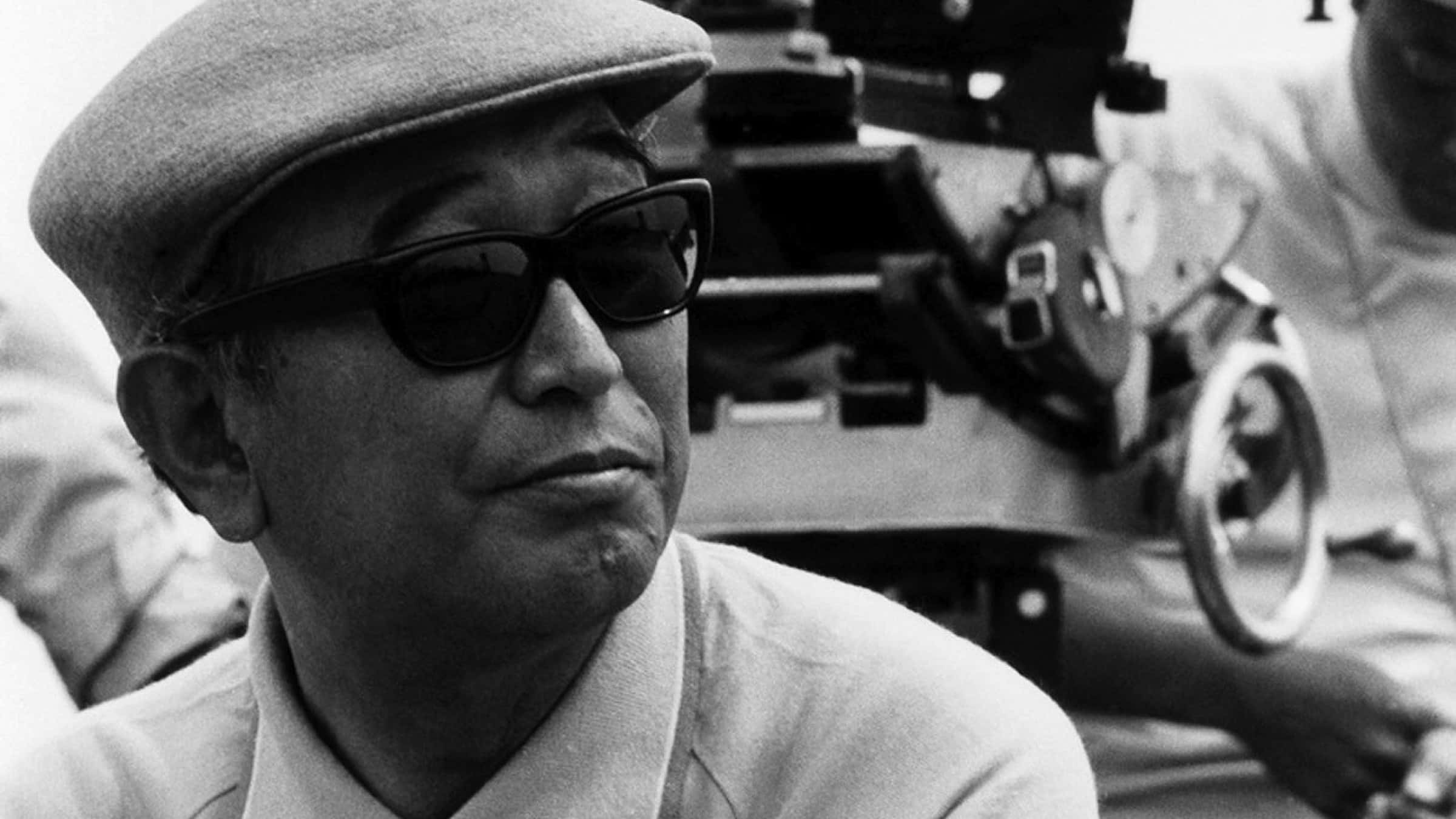 Akira Kurosawa 桌布 壁紙 — 一個戴帽子和太陽眼鏡的男人的黑白肖像，背景有一部底片相機。