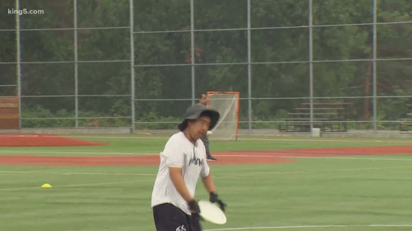 1366x768 Ultimate Frisbee 桌布 壁紙 — 一個戴帽子和手套的人準備在綠色運動場上擲飛盤，背景有紅色跑道和鐵絲網圍欄。