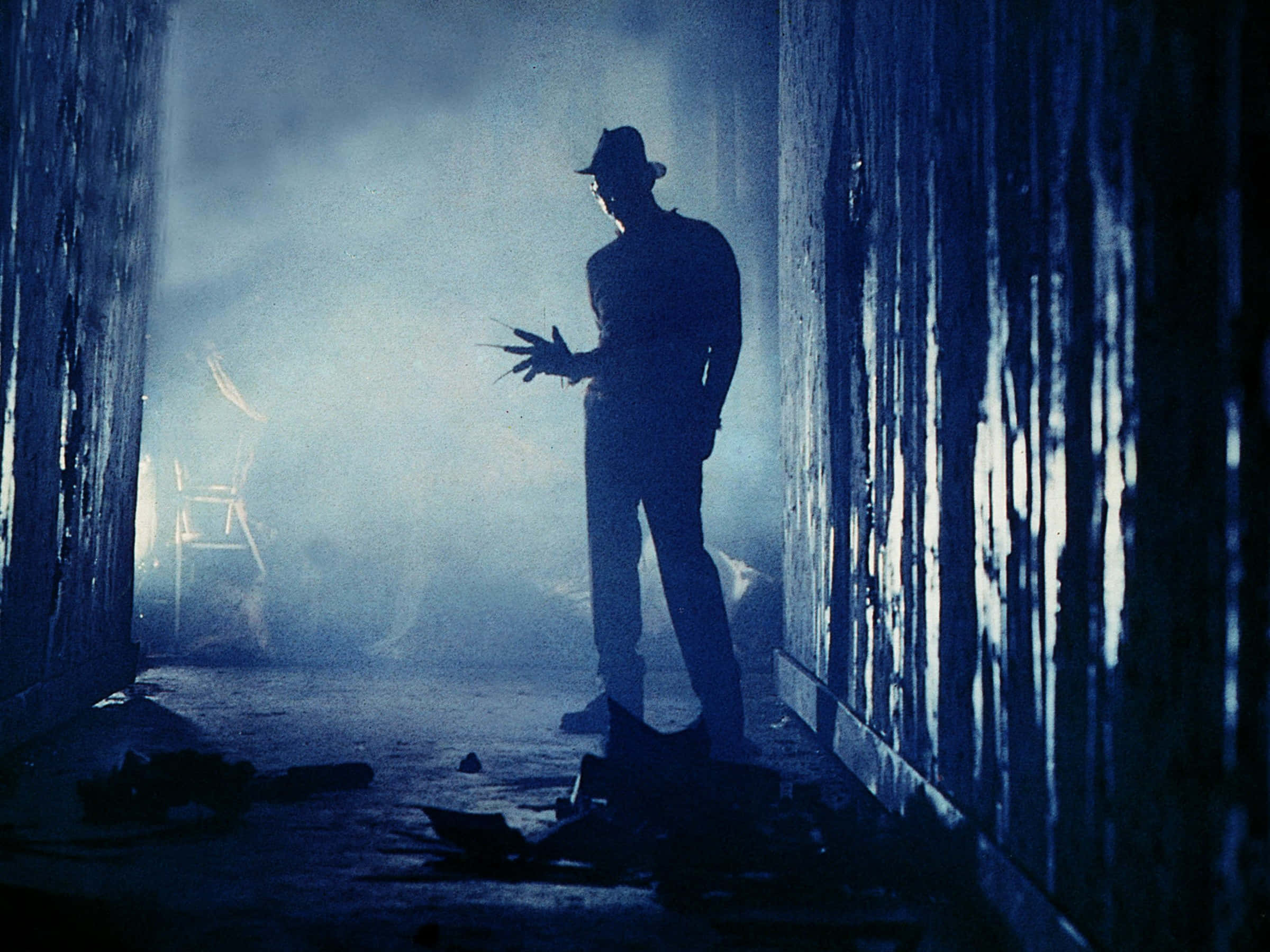 Freddy Krueger 桌布 壁紙 — 一個戴帽子和手套的男人的威脅性剪影，站在霧氣瀰漫、藍色燈光的走廊中，背景有碎片和椅子。