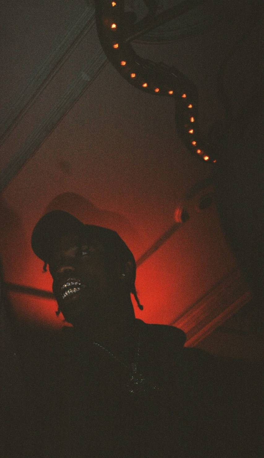 Travis Scott Aesthetic 桌布 壁紙 — 一個戴帽子和燒烤架的剪影人物在昏暗的房間裡，在溫暖的紅色燈串下微笑。