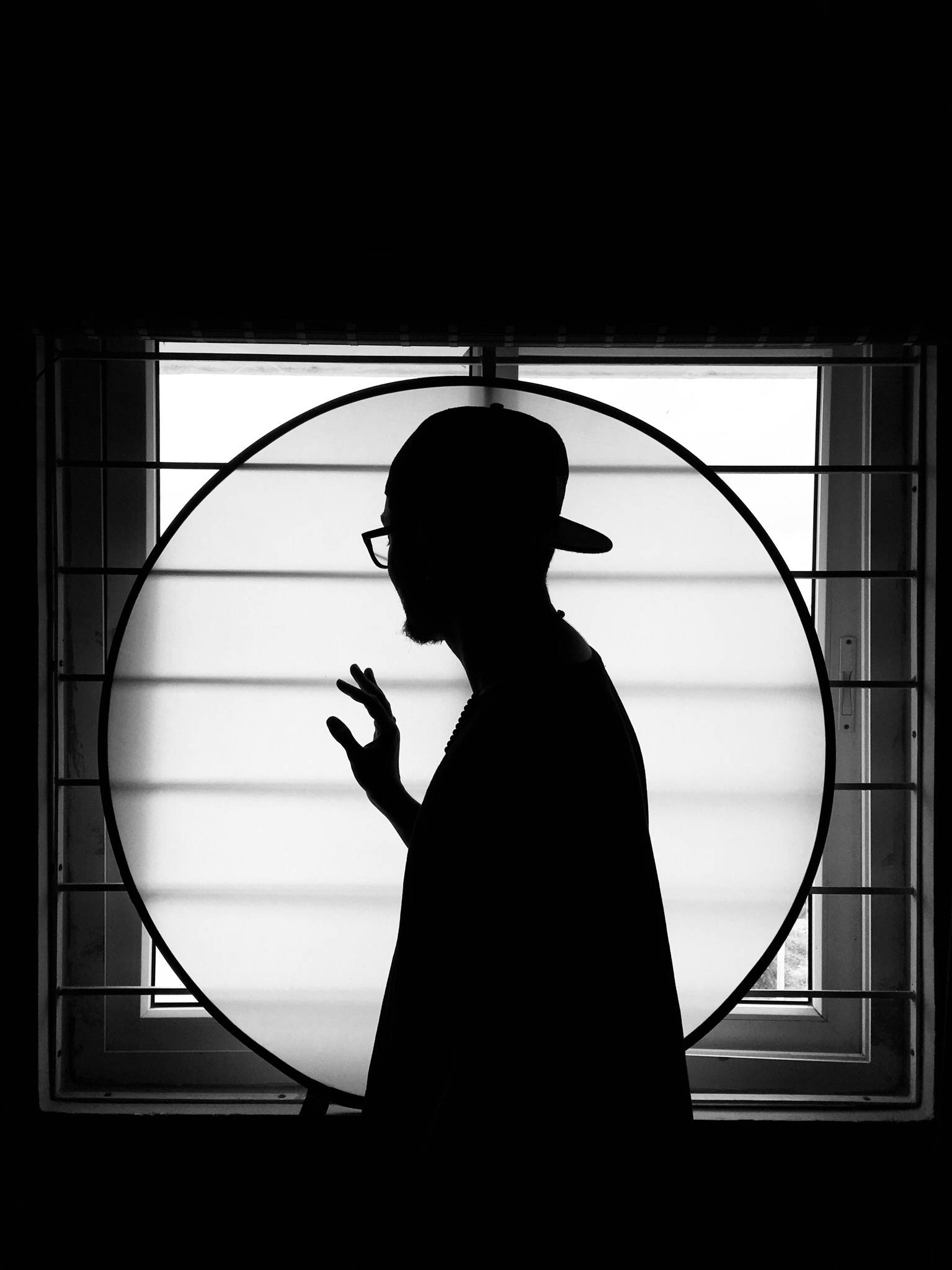 黑白高畫質 桌布 壁紙 — 一個戴帽子和眼鏡的男人的鮮明黑白剪影，被一個圓形窗戶框住，背景明亮。
