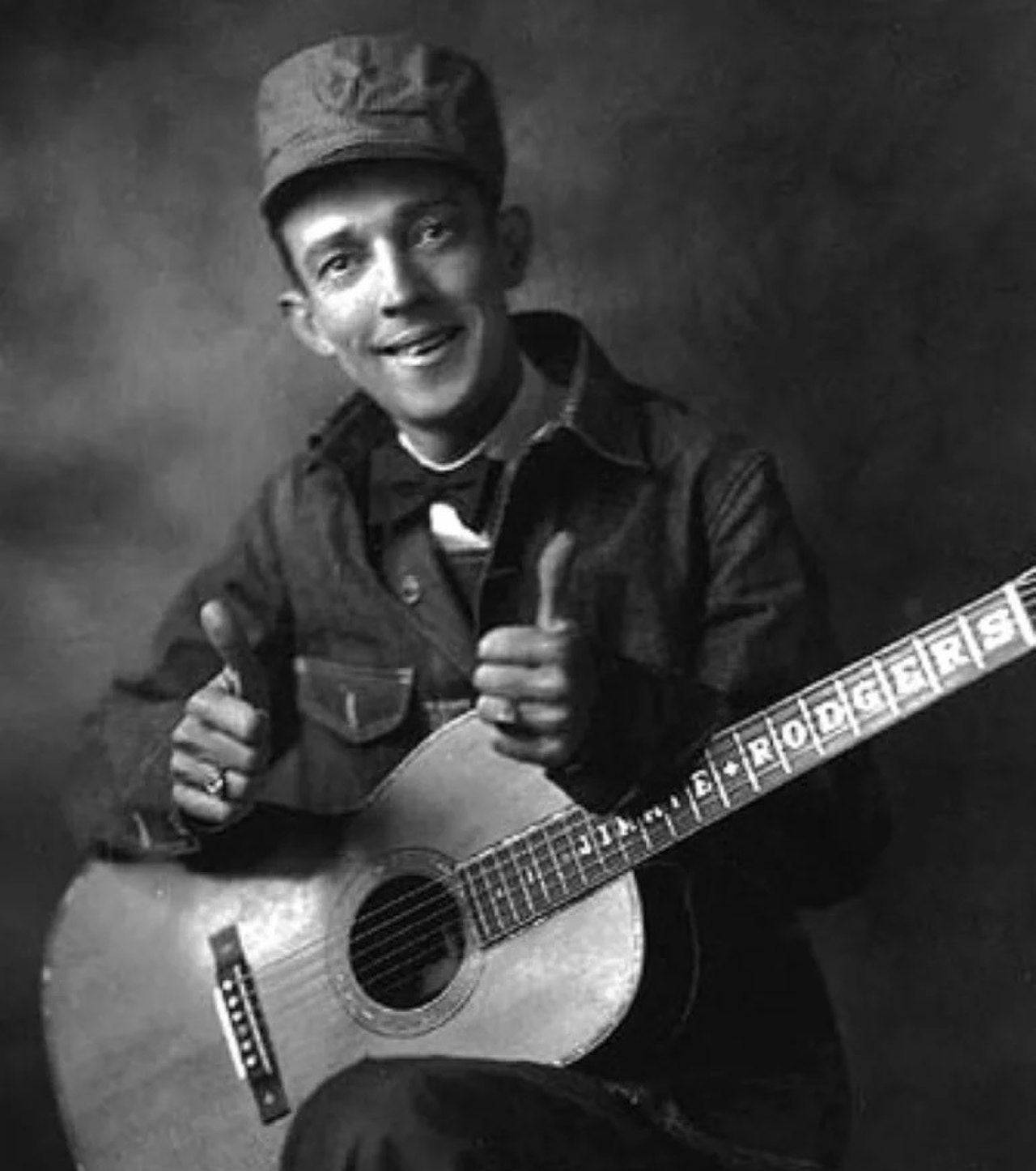 guitar player 桌布 壁紙 — 一個戴帽微笑的男人豎起兩個大拇指，同時抱著一把指板上有他名字的木吉他。
