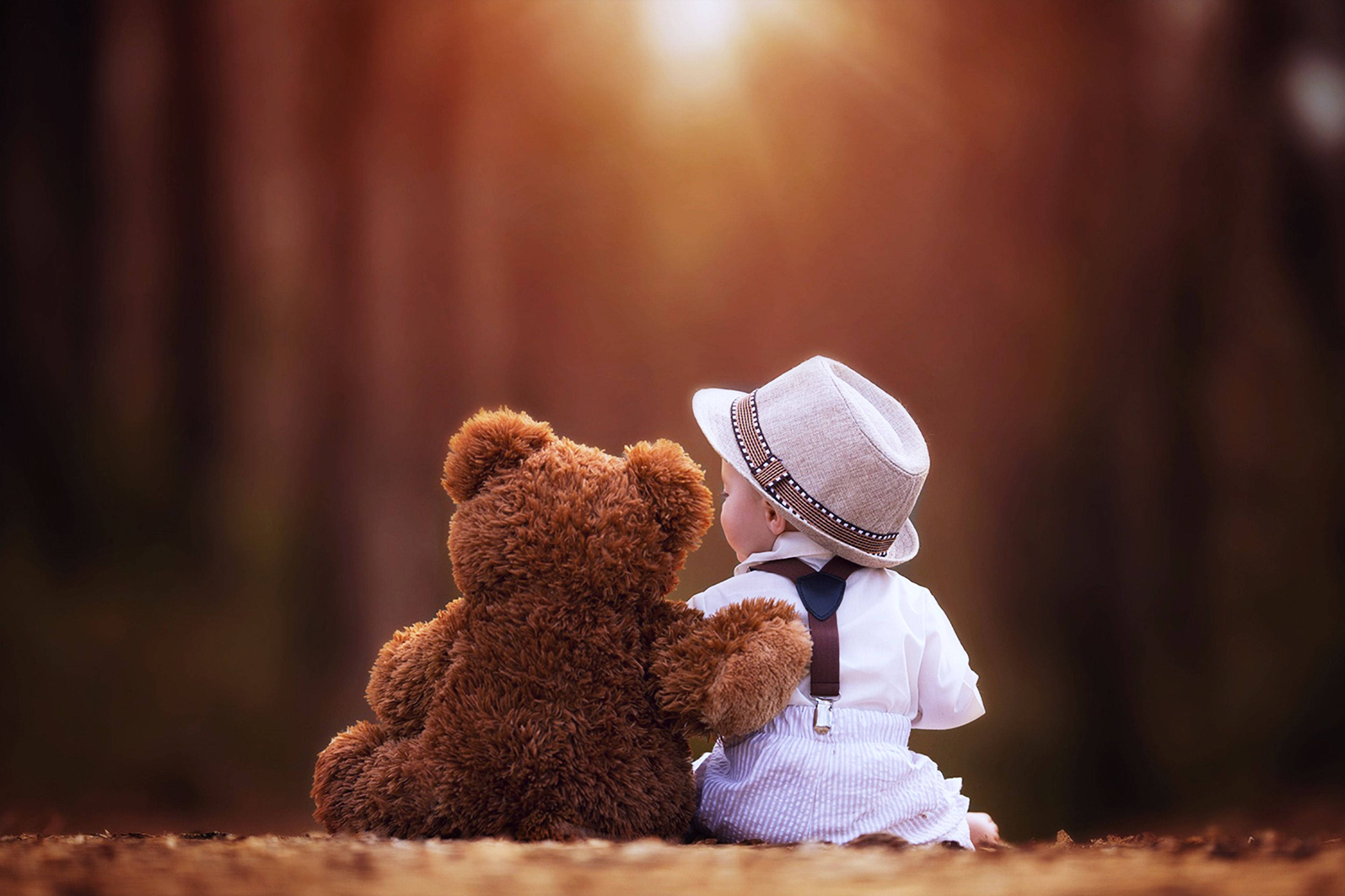 Cute Bear 桌布 壁紙 — 一個戴帽的幼兒坐在棕色泰迪熊旁邊，凝視著穿過森林的金色陽光。