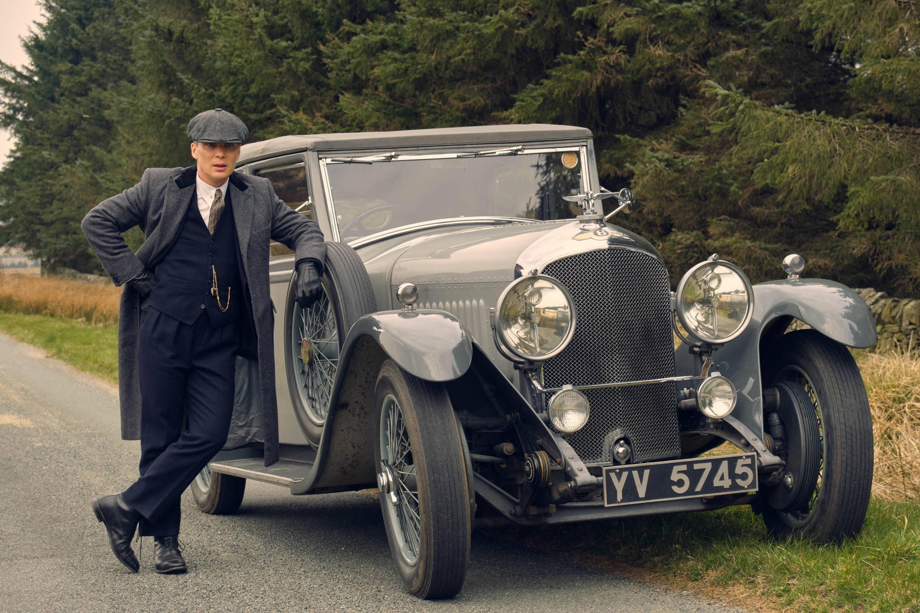 Thomas Shelby 桌布 壁紙 — 一個戴平頂帽、穿西裝的男人靠在一輛停在鄉村路邊、旁邊有常綠樹的復古銀色汽車上。