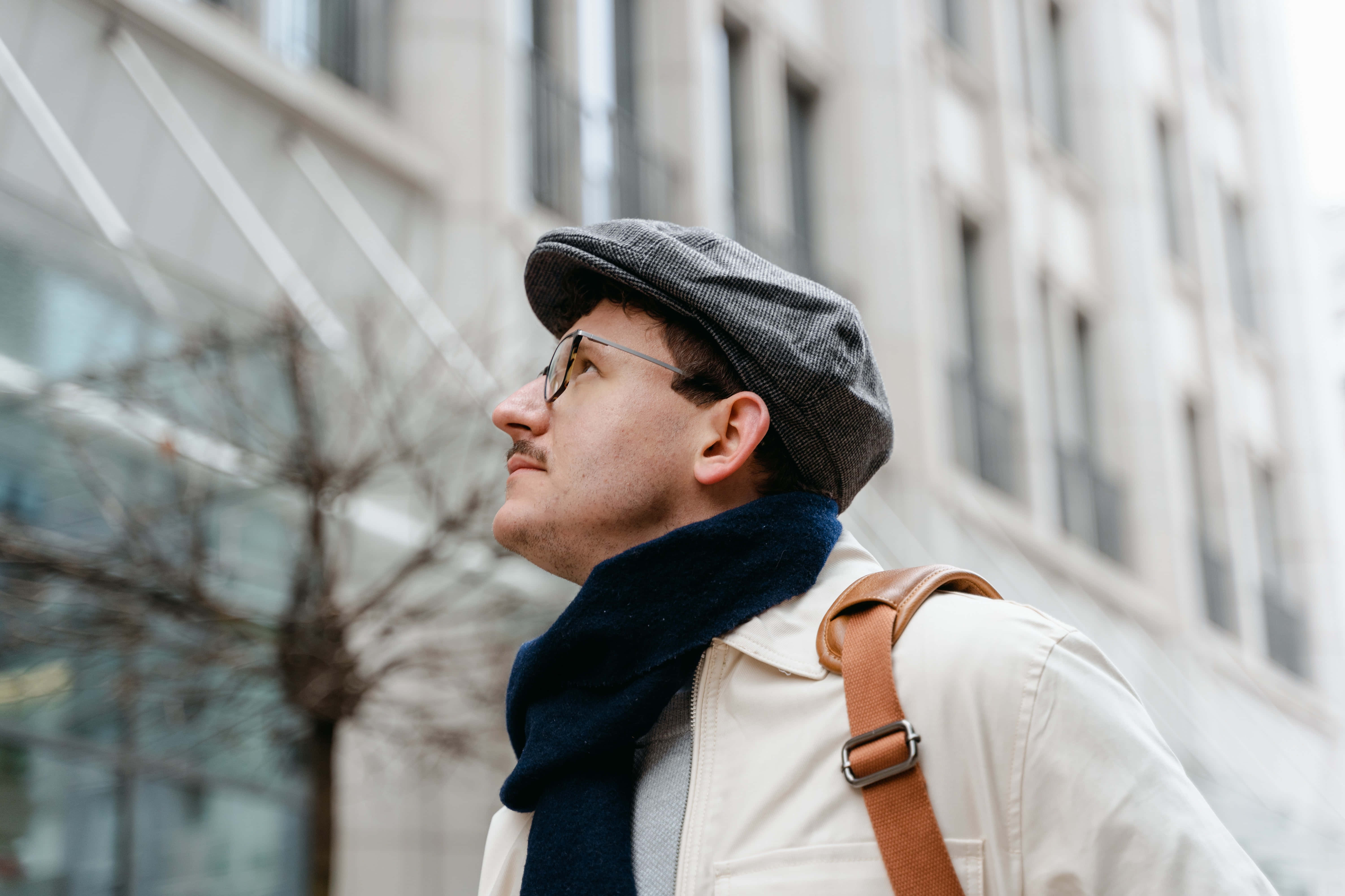 Navy Blue Scarf 桌布 壁紙 — 一個戴平頂帽和圍巾的男人在一個有光禿樹木和現代建築物在後面的城市街道上向上凝視。