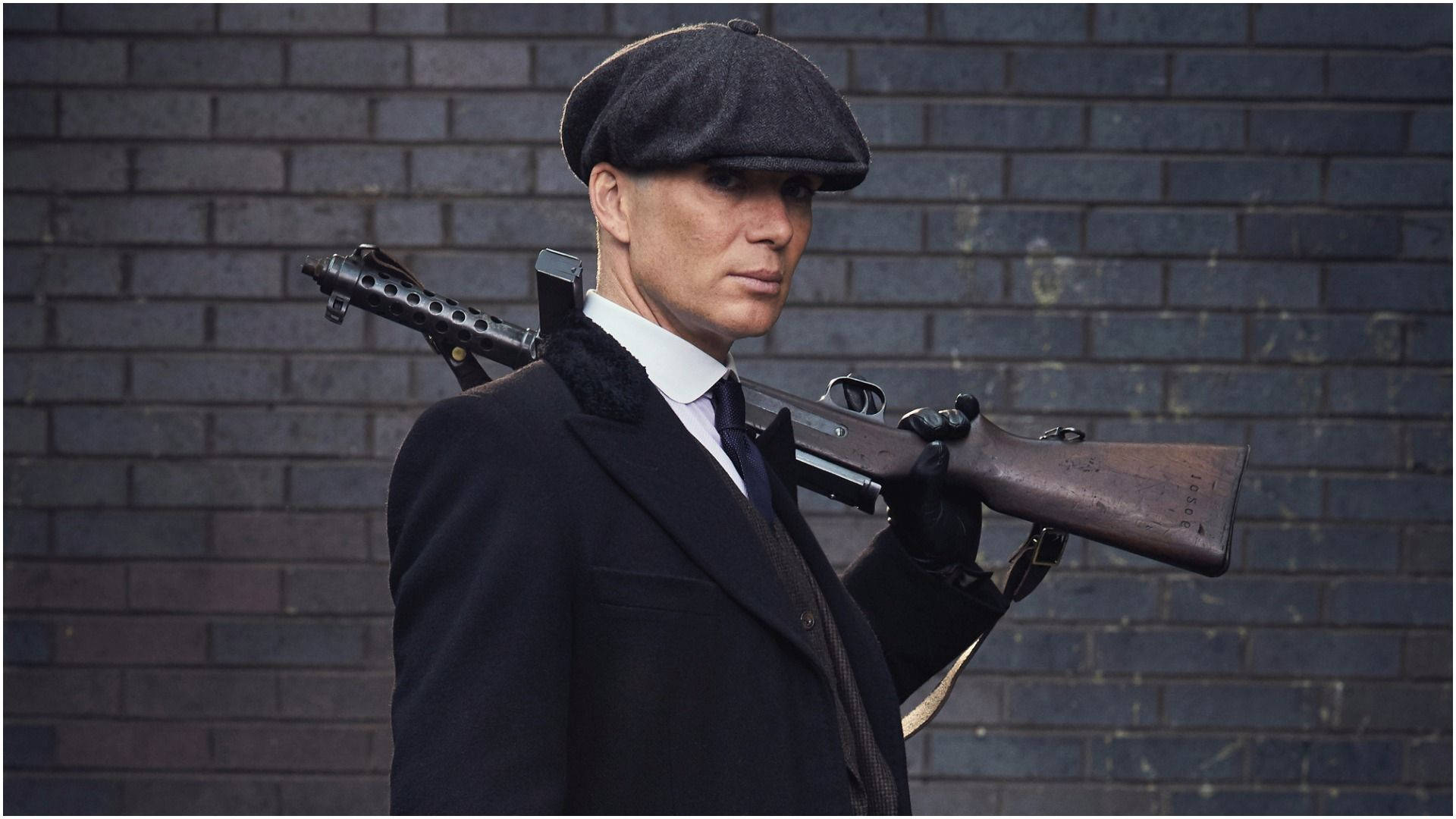戴平頂帽和穿風衣的男人手持復古衝鋒槍靠著磚牆。 — Peaky Blinders 壁紙