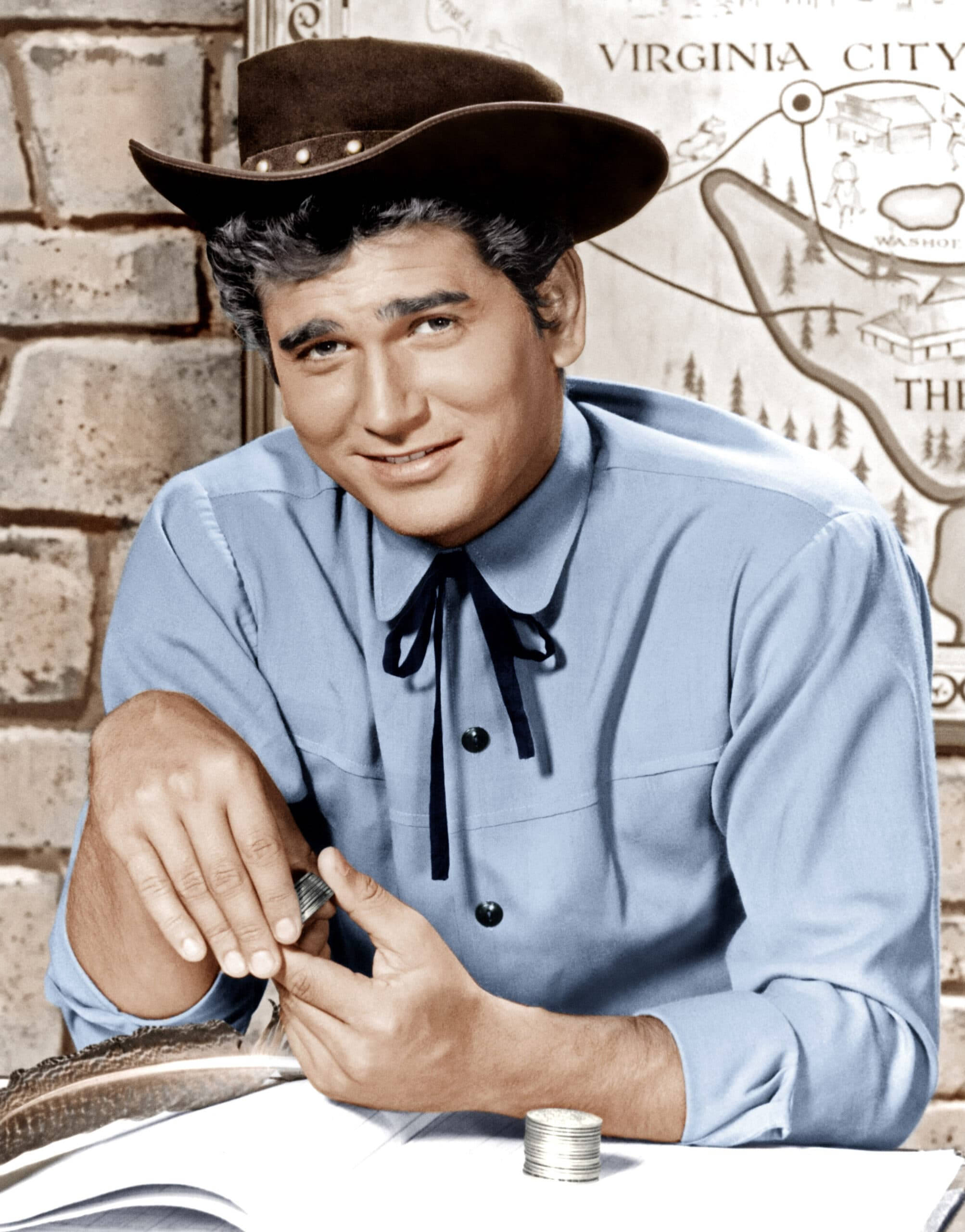 Michael Landon 桌布 壁紙 — 一個戴棕色帽、穿藍色襯衫的微笑牛仔，靠在一張有硬幣和羽毛筆的桌子上，背景是磚牆和地圖。