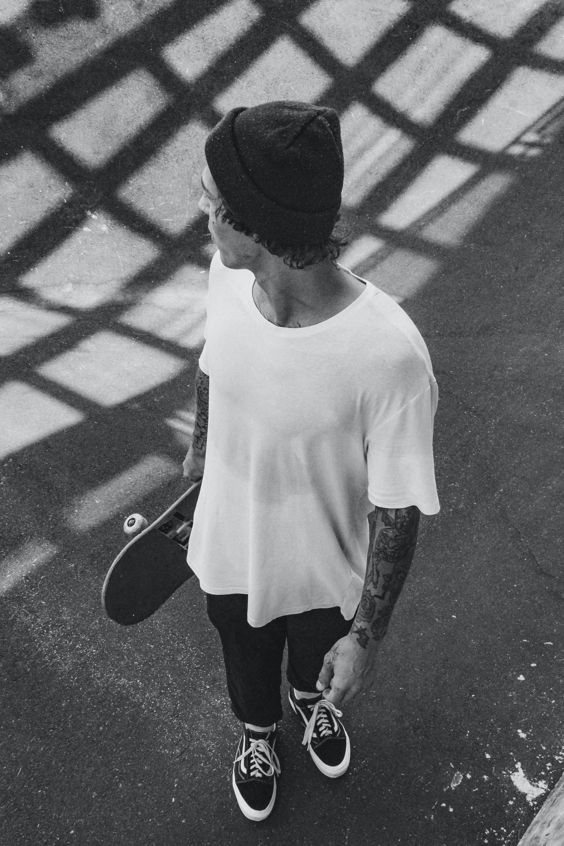 Skater Boy 桌布 壁紙 — 一個戴毛帽、有刺青的年輕男子，在人行道的陰影網格下拿著滑板。