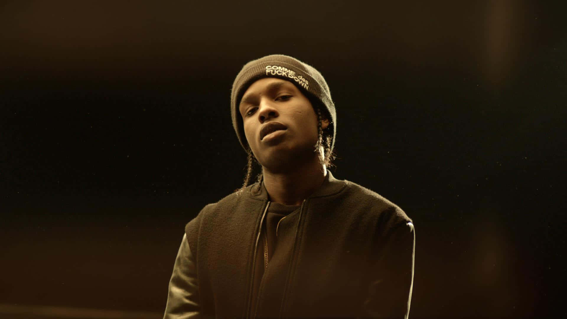 ASAP Rocky 桌布 壁紙 — 一個戴毛帽和夾克的男人站在昏暗的金色光線中，背景黑暗，有漂浮的粒子。