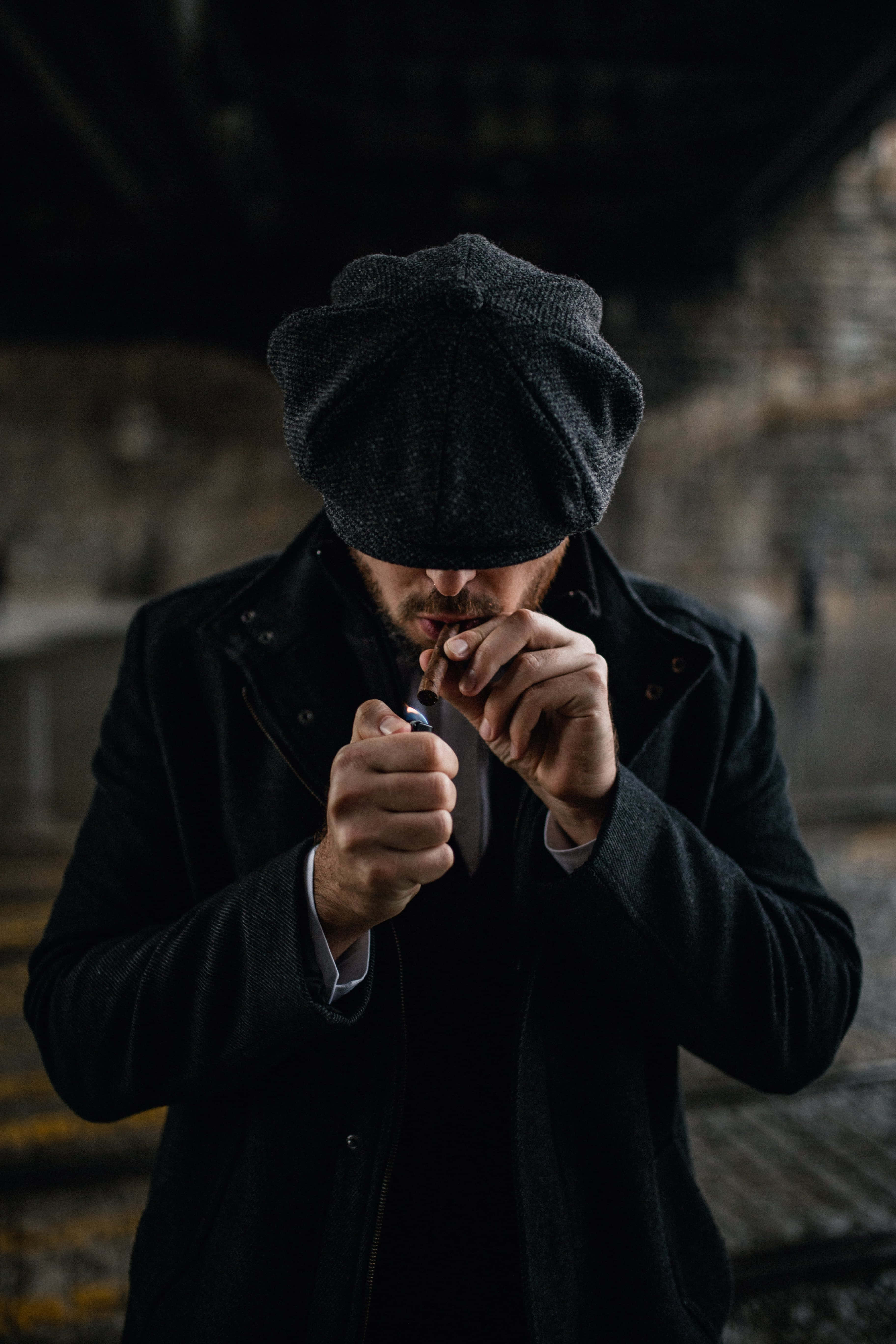 Black Gangster 桌布 壁紙 — 一個戴深色平頂帽、穿深色外套的男人，在昏暗骯髒的城市天橋下點煙。