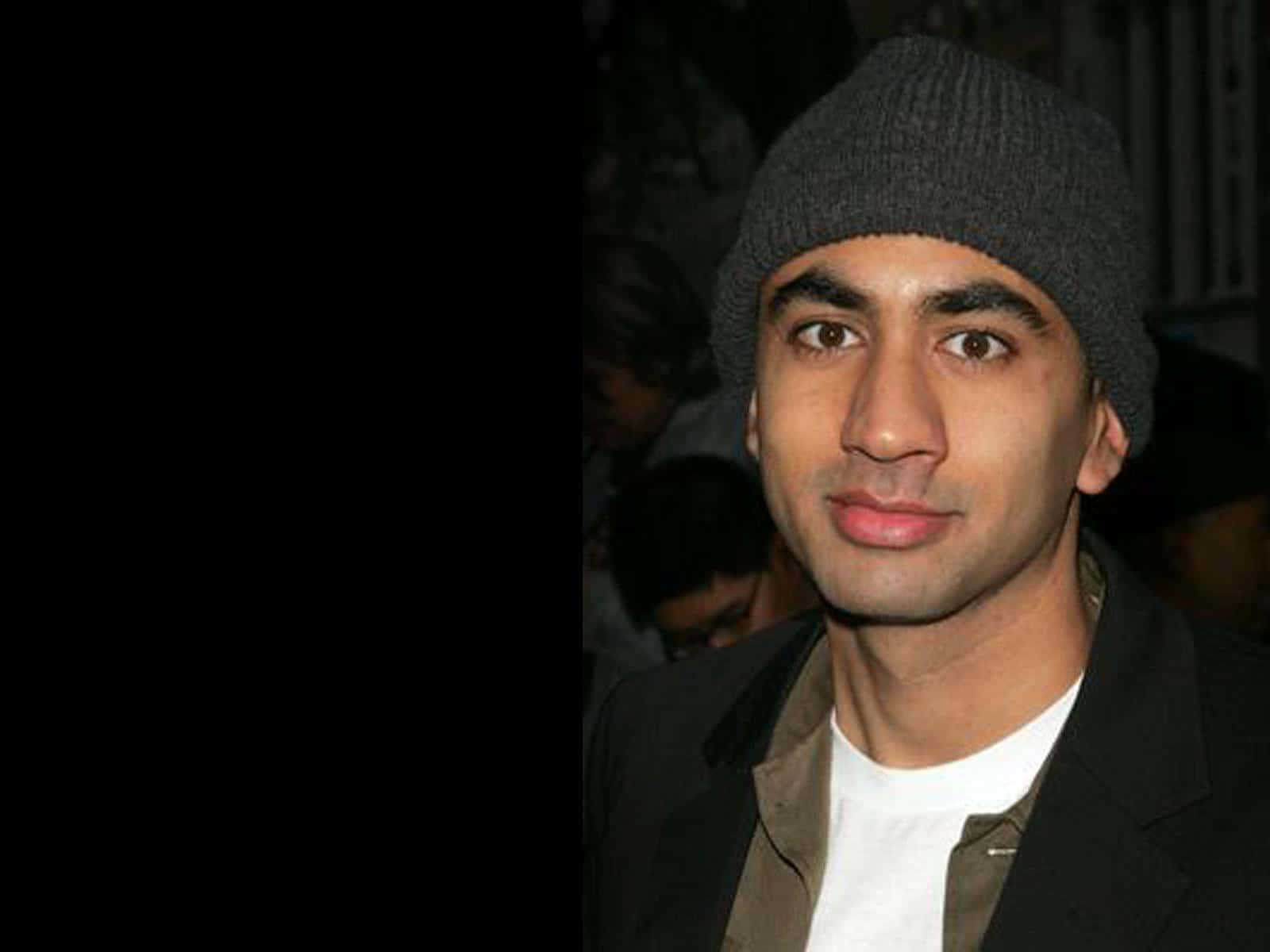 Kal Penn 桌布 壁紙 — 一個戴灰色毛帽、穿深色外套的男人，在昏暗的人群背景下輕輕微笑。