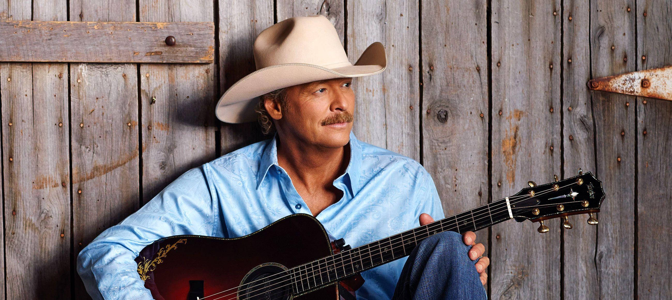 Alan Jackson 桌布 壁紙 — 一個戴牛仔帽、穿藍色襯衫的男人靠著風化木頭，手持木吉他。
