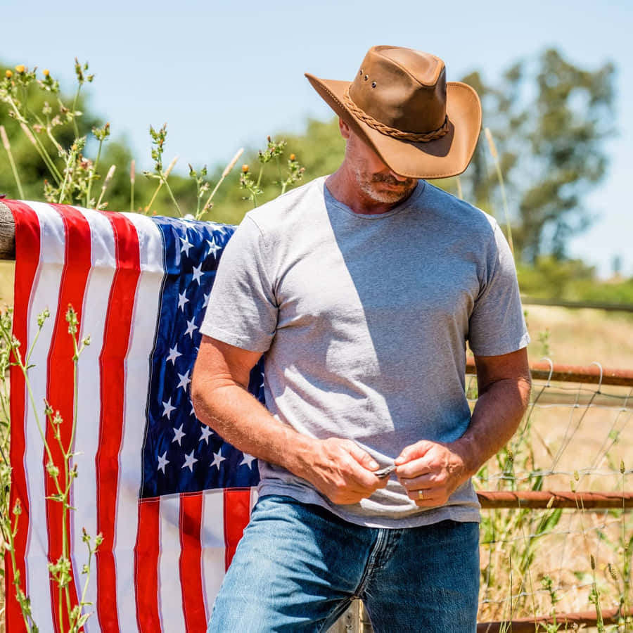 Cowboy Hat 桌布 壁紙 — 一個戴牛仔帽的男人站在籬笆旁，籬笆上披著美國國旗，在明亮的陽光下。
