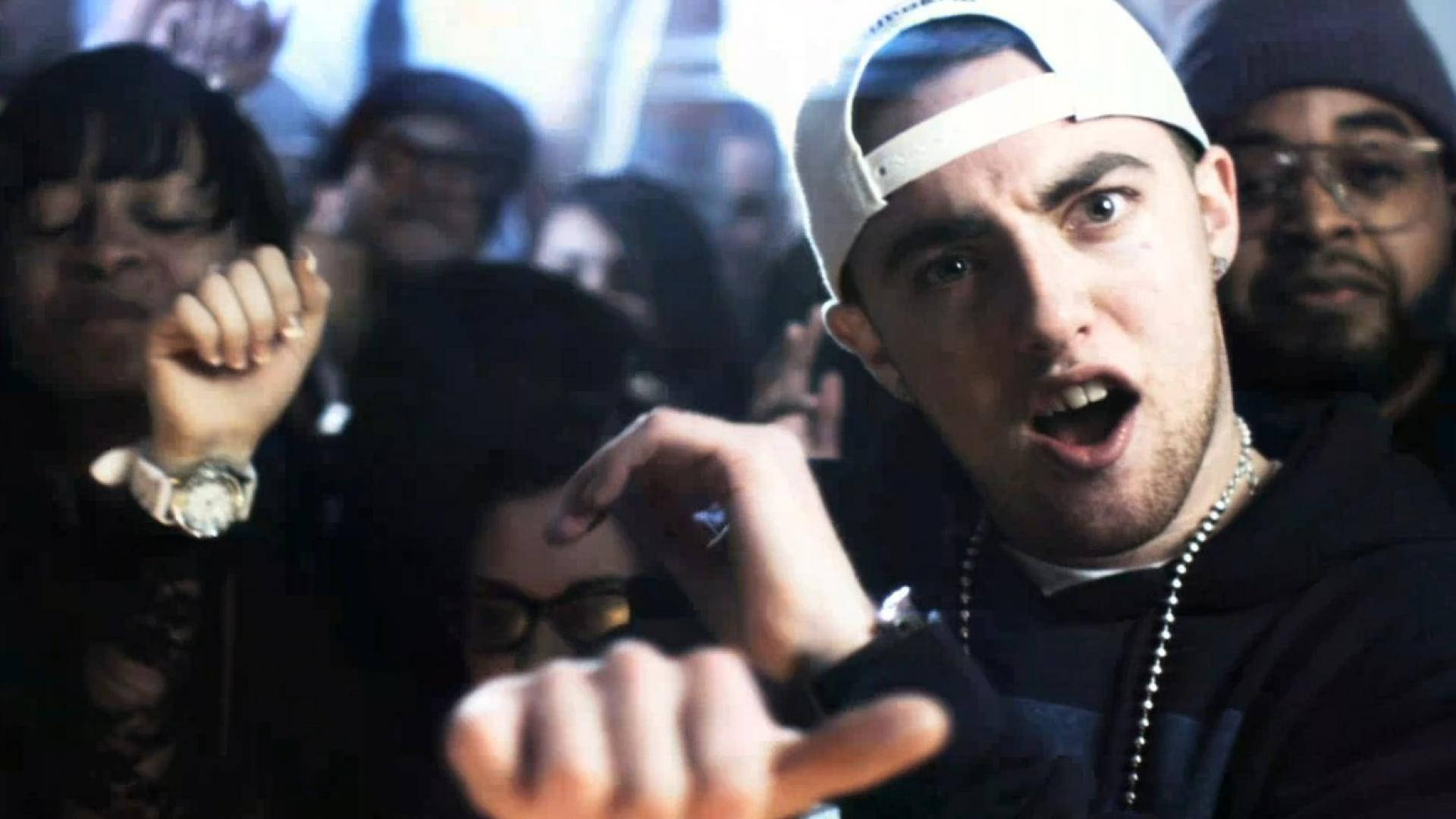 Mac Miller 桌布 壁紙 — 一個戴白色帽子的年輕人在昏暗的城市環境中，被人群包圍，強烈地向前指。