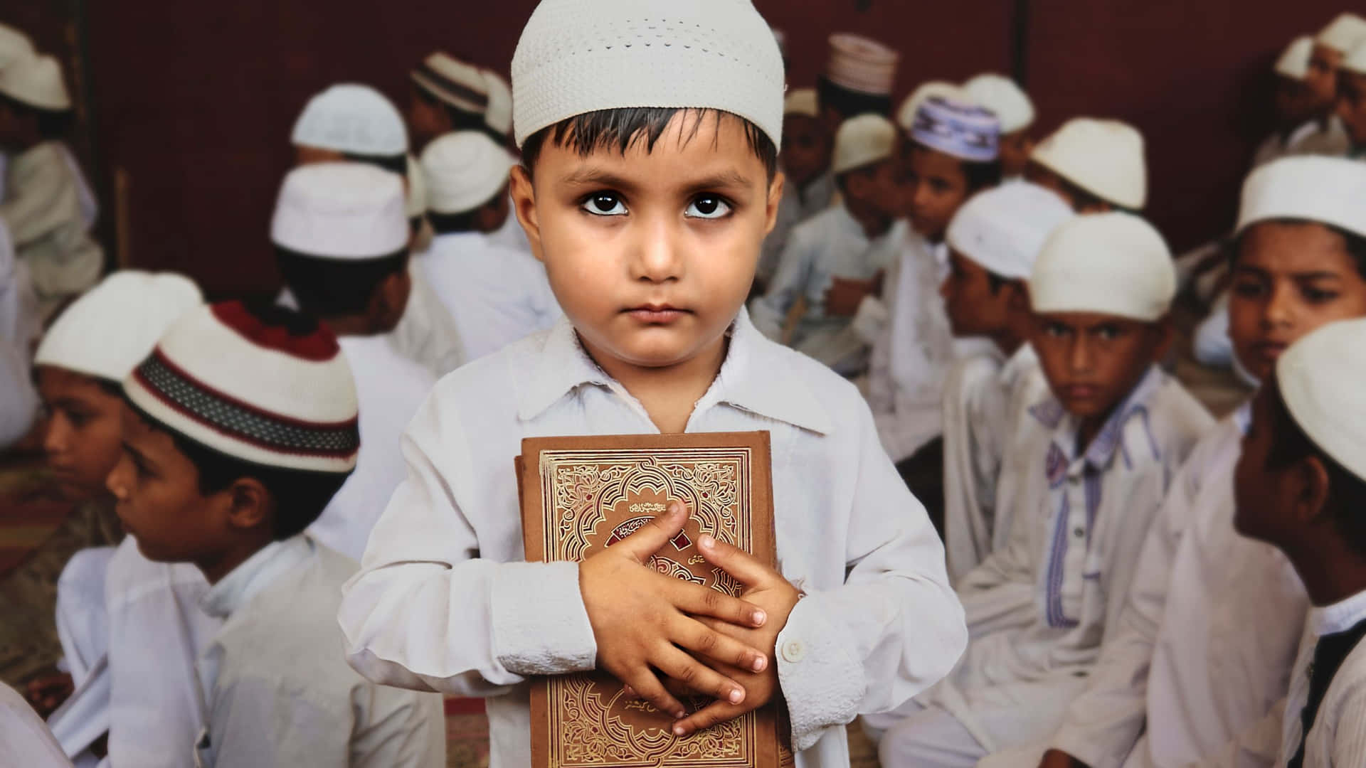 muslim 桌布 壁紙 — 一個戴白色帽子的年輕男孩在宗教聚會中與其他孩子坐在一起，手持古蘭經。