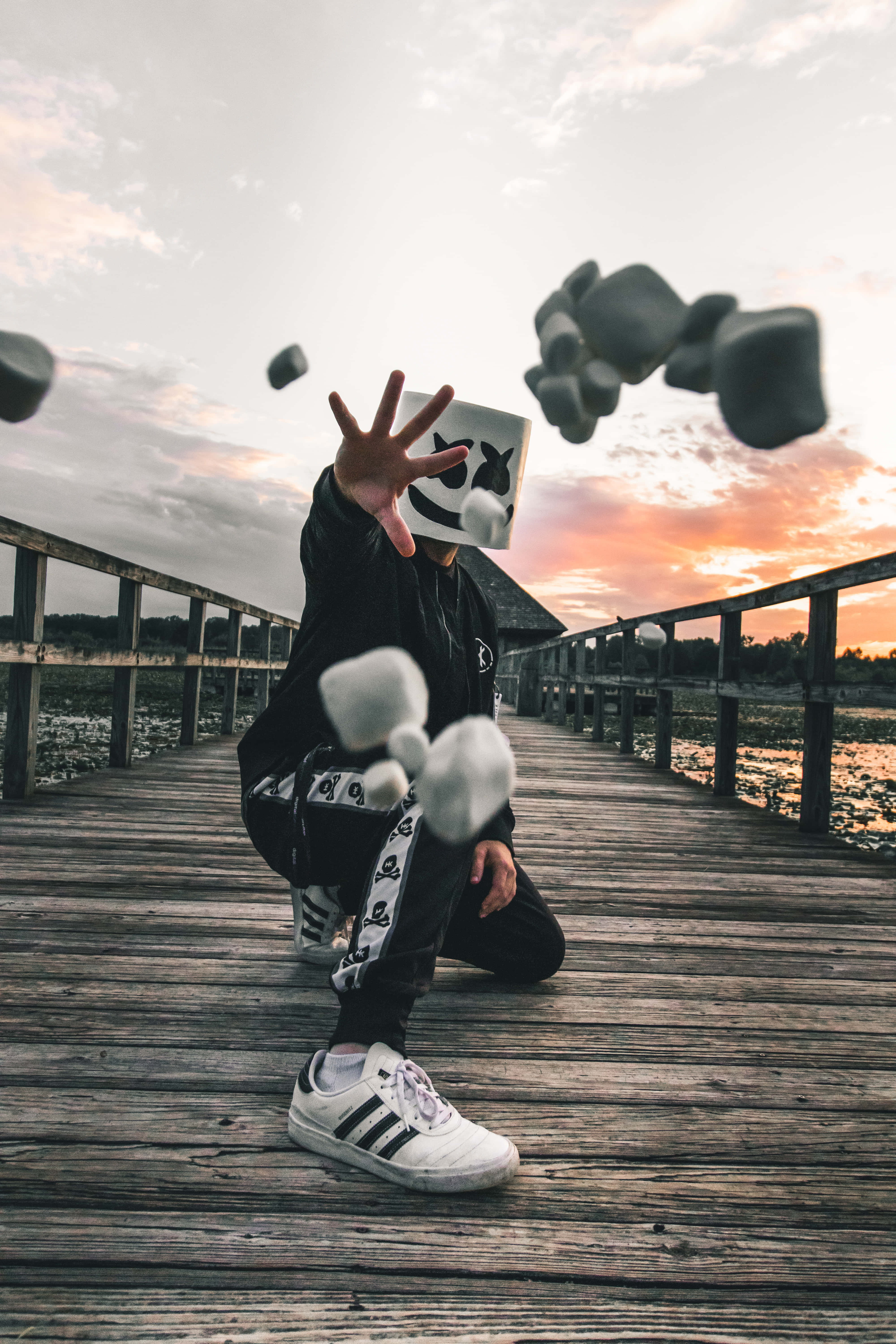 Marshmello 動態圖 桌布 壁紙 — 一個戴白色面具、穿黑色衣服的人蹲在木碼頭上，在日落時將石頭拋向空中。