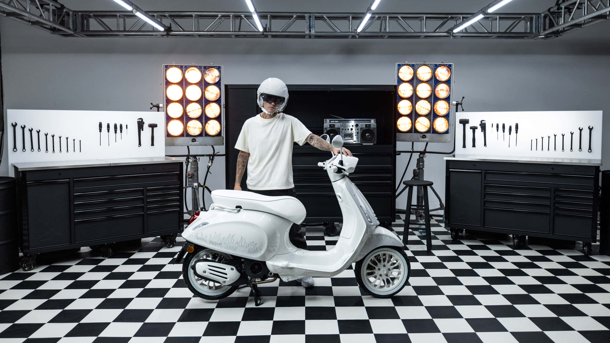 scooter 桌布 壁紙 — 一個戴白色頭盔的人站在一輛白色機車旁，身處高對比度、地板呈棋盤格並展示工具的車庫中。