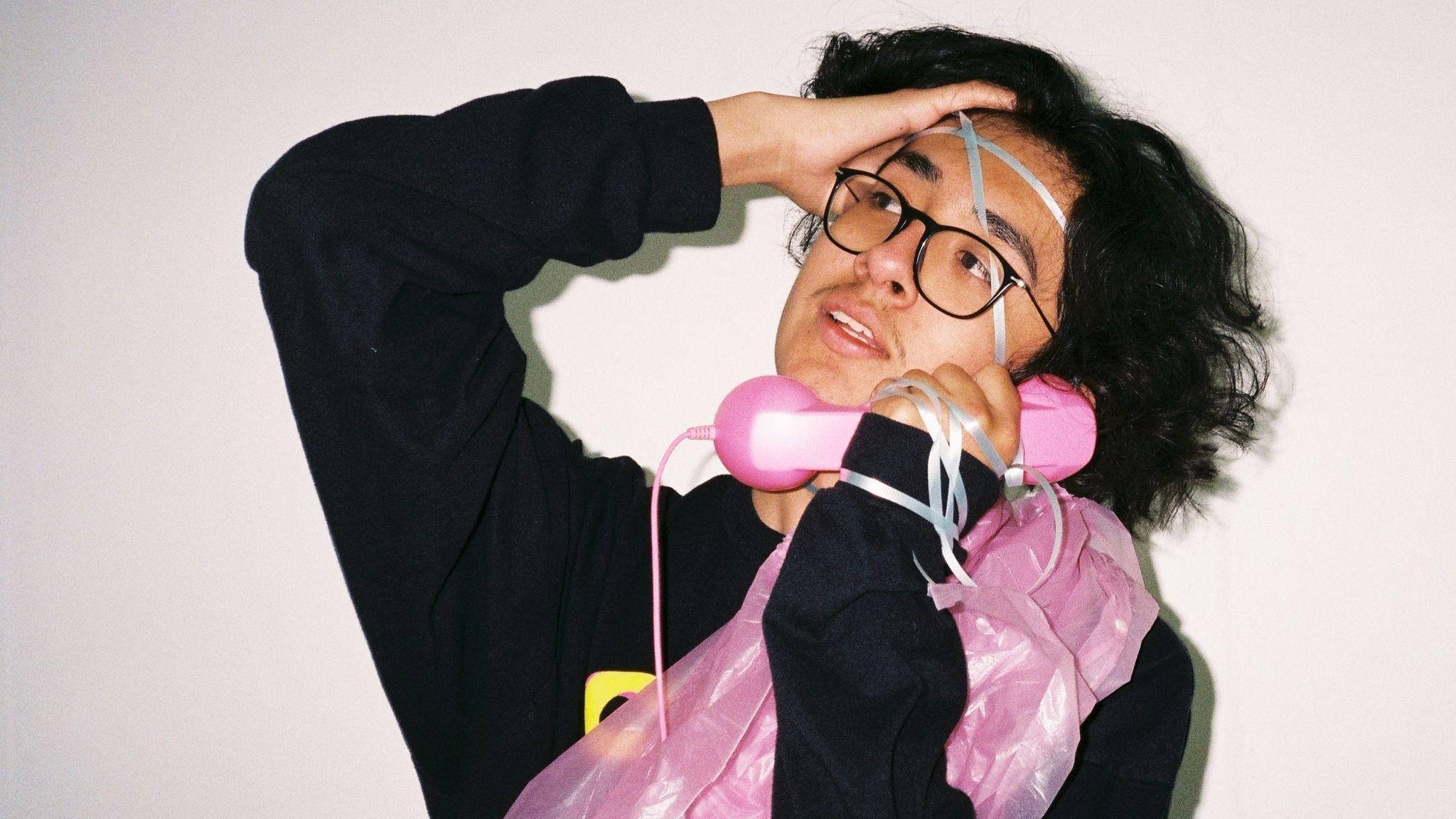 Cuco 桌布 壁紙 — 一個戴眼鏡、深色頭髮的人，在純白牆壁前，拿著一個被絲帶纏繞的粉紅色電話聽筒。