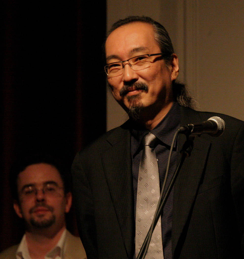 satoshi kon 桌布 壁紙 — 一個戴眼鏡、留鬍子、穿西裝的男人在燈光昏暗的舞台上微笑著對著麥克風說話。