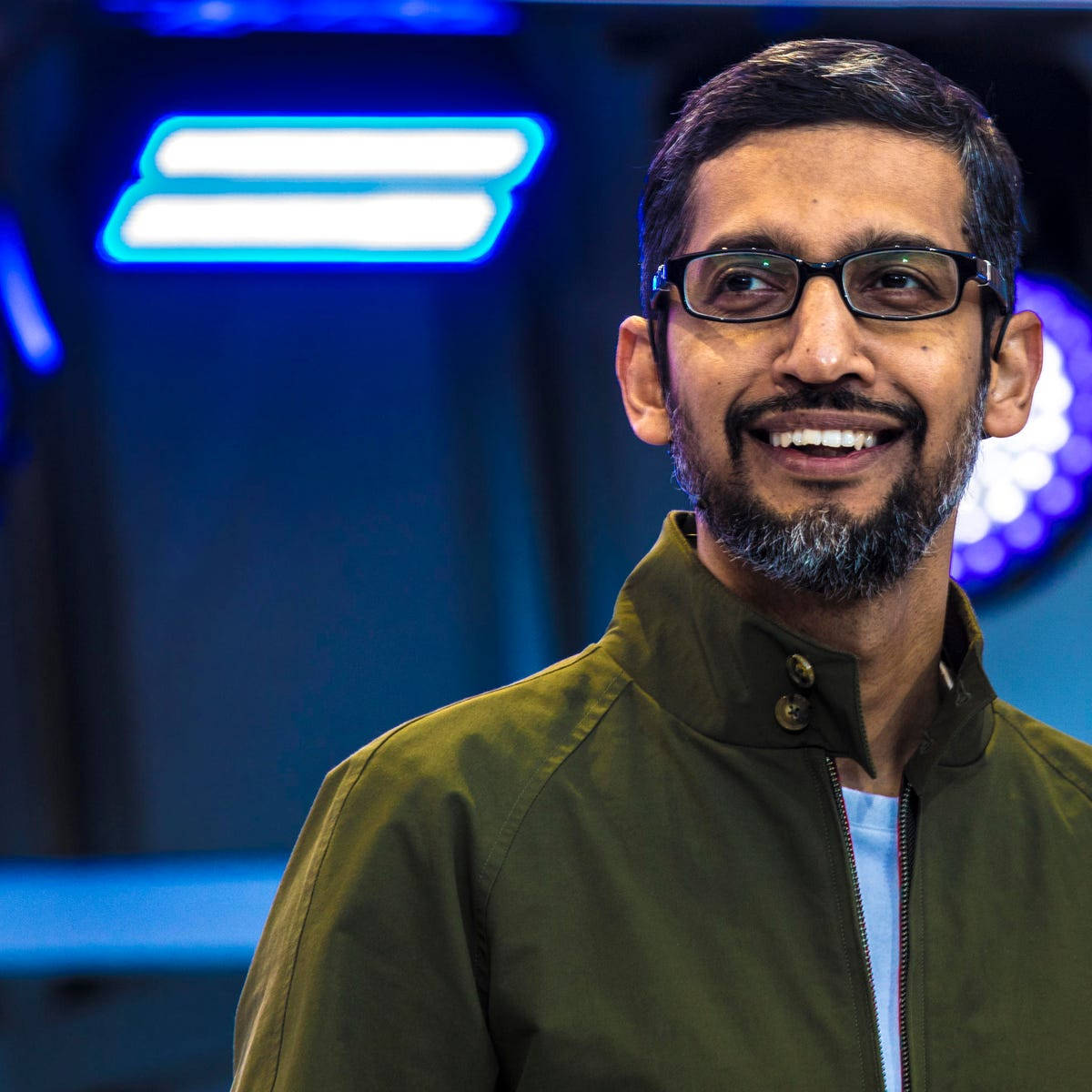 google 桌布 壁紙 — 一個戴眼鏡、留鬍鬚、面帶微笑的男人，身穿橄欖色外套，背景是藍色霓虹燈光。