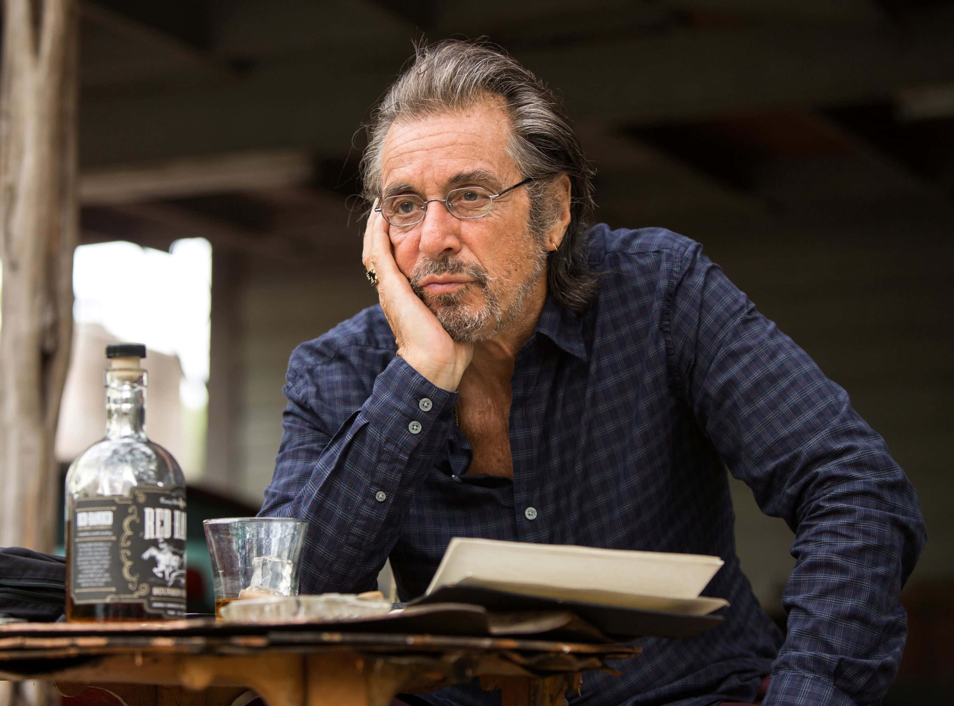 Al Pacino 桌布 壁紙 — 一個戴眼鏡、留鬍鬚的男人，在一個質樸的木桌旁，旁邊放著一瓶威士忌和一個杯子，他用手托著頭休息。