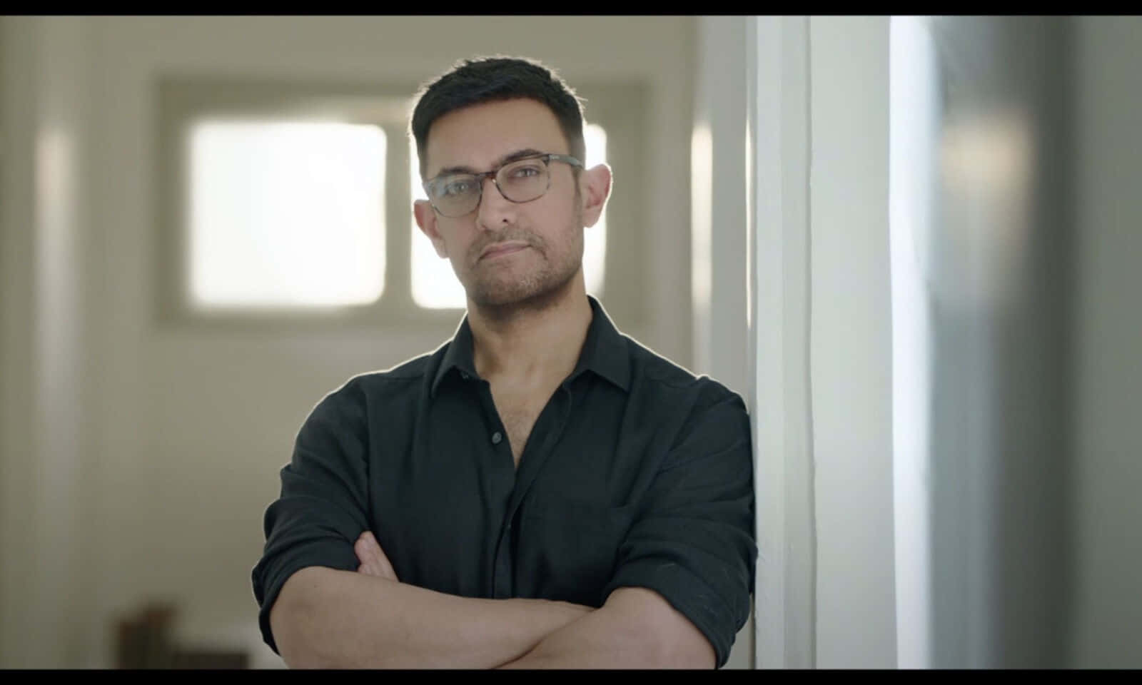 Aamir Khan 桌布 壁紙 — 一個戴眼鏡、穿黑色襯衫的男人自信地交叉雙臂，站在光線柔和的室內走廊。