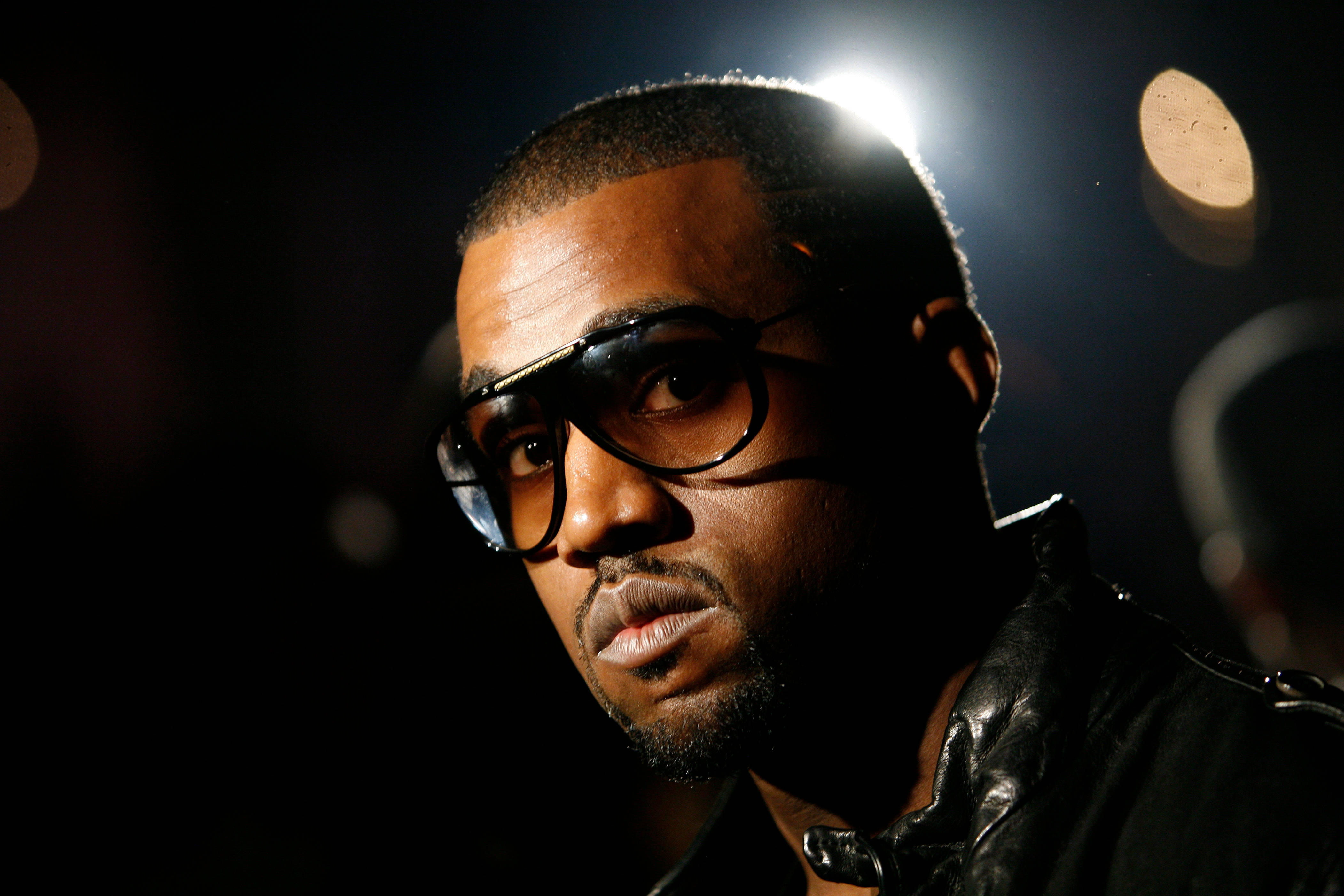 Kanye 桌布 壁紙 — 一個戴眼鏡、身穿皮夾克的男人，在戲劇性的聚光燈下，對著一個有散景燈光的黑暗背景，用力凝視。
