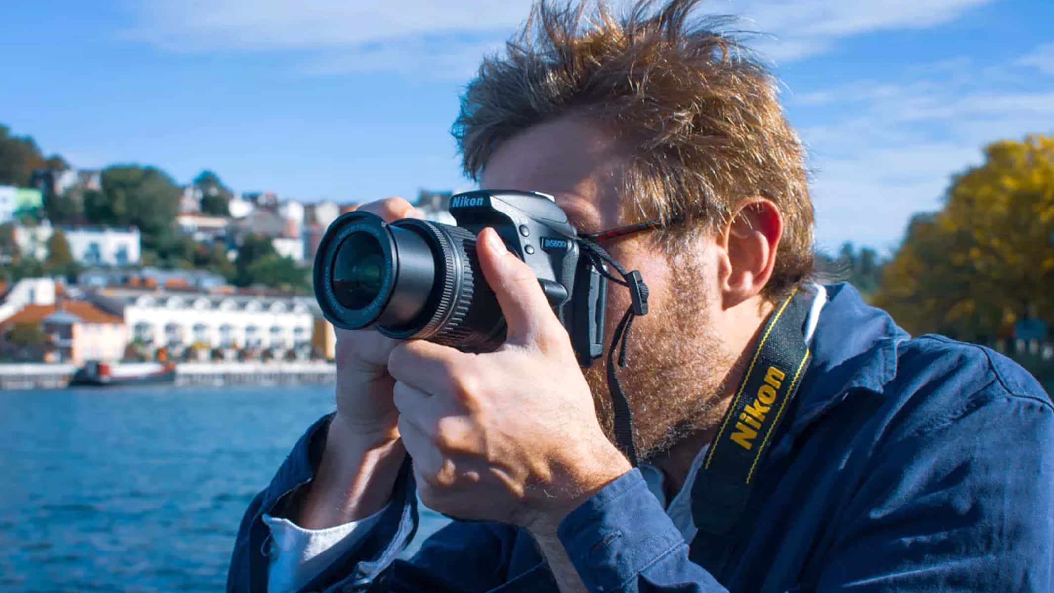 Dslr Camera 桌布 壁紙 — 一個戴眼鏡、身穿藍色外套的男人，在晴朗的藍天下，於海濱小鎮用 Nikon DSLR 相機拍照。
