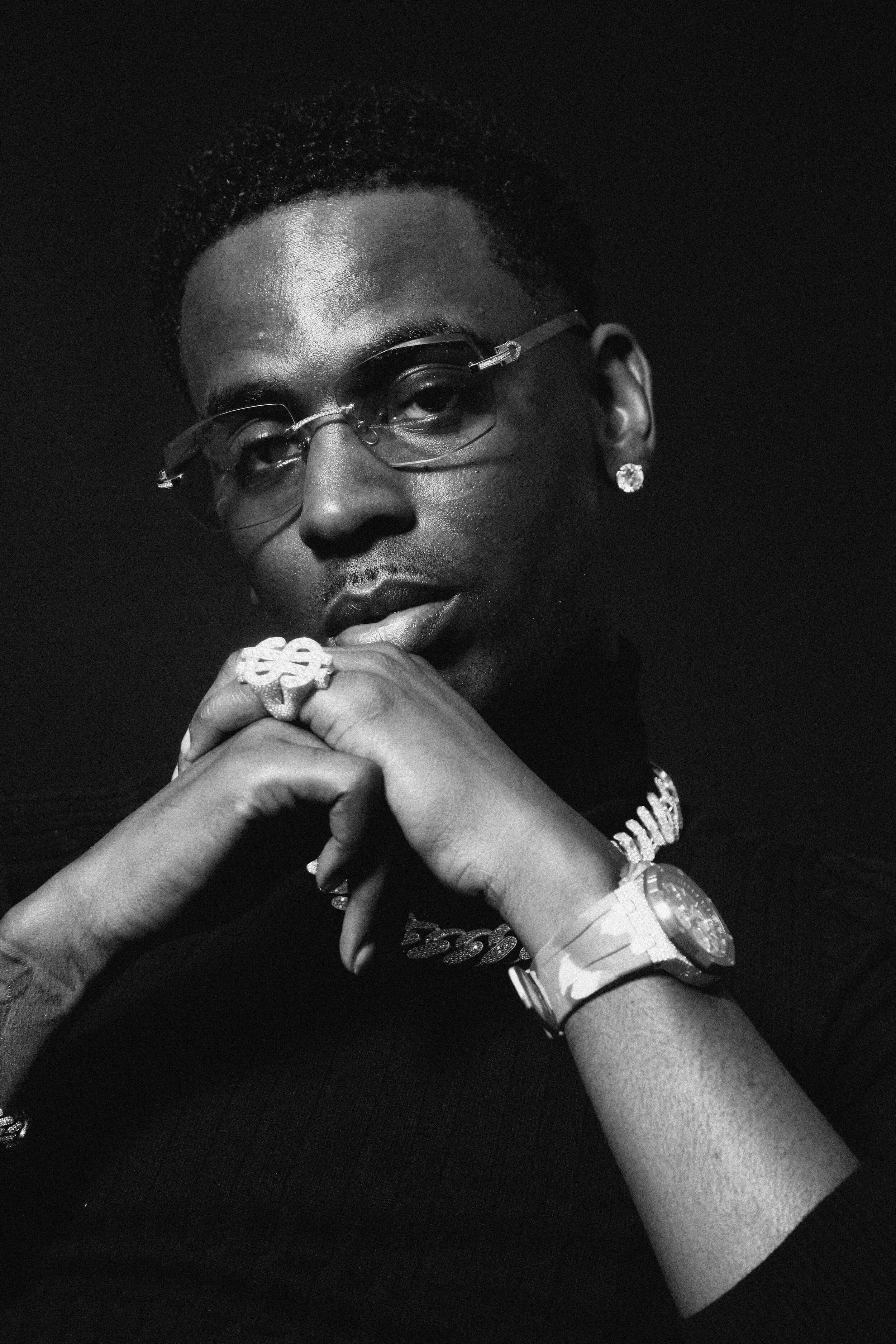 Young Dolph 桌布 壁紙 — 一個戴眼鏡和珠寶的男人，雙手交叉放在下巴下的黑白肖像。