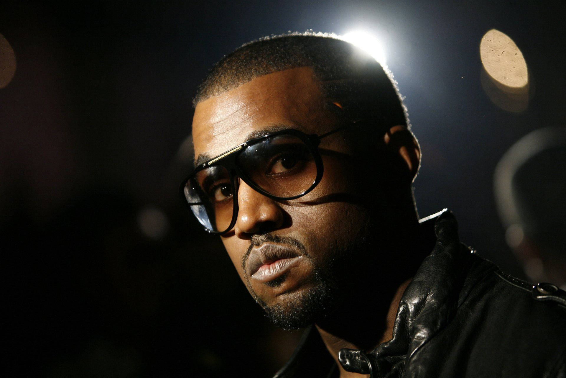 Kanye West 桌布 壁紙 — 一個戴眼鏡和皮夾克的男人在戲劇性舞台燈光下，背景有散景效果，他凝視著。