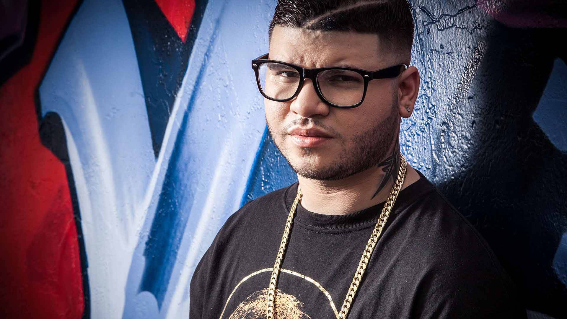 farruko 桌布 壁紙 — 一個戴眼鏡和金色鏈條的男人自信地站在一面充滿活力的塗鴉牆前，牆上有大膽的紅色、藍色和黑色。