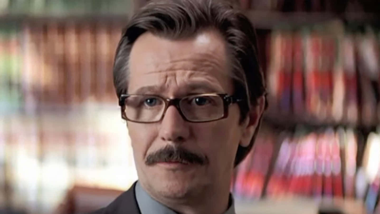 Commissioner Gordon 桌布 壁紙 — 一個戴眼鏡和鬍子的男人在溫暖燈光的圖書館中專注地凝視，身後是模糊的書架。