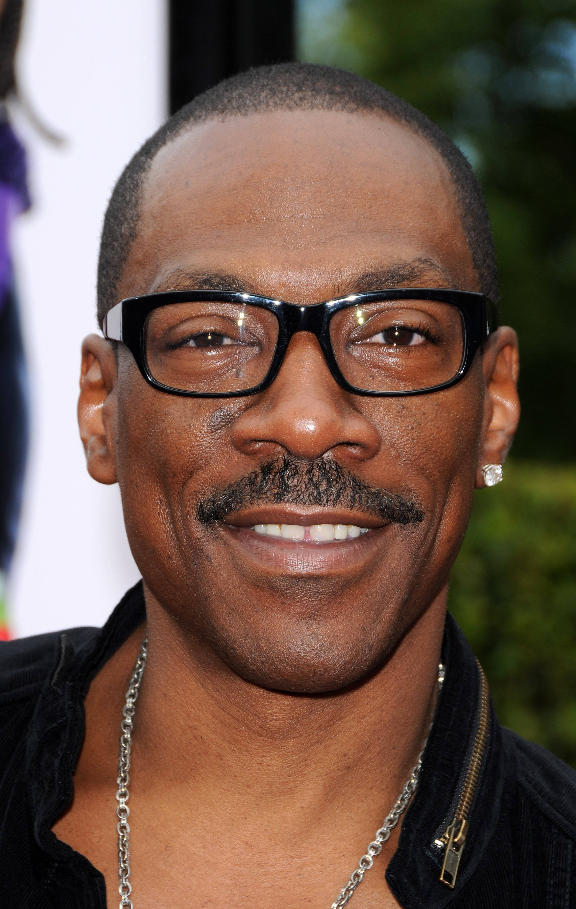 eddie murphy 桌布 壁紙 — 一個戴眼鏡和鬍鬚、穿著黑色外套和銀鏈的微笑男子的特寫肖像。